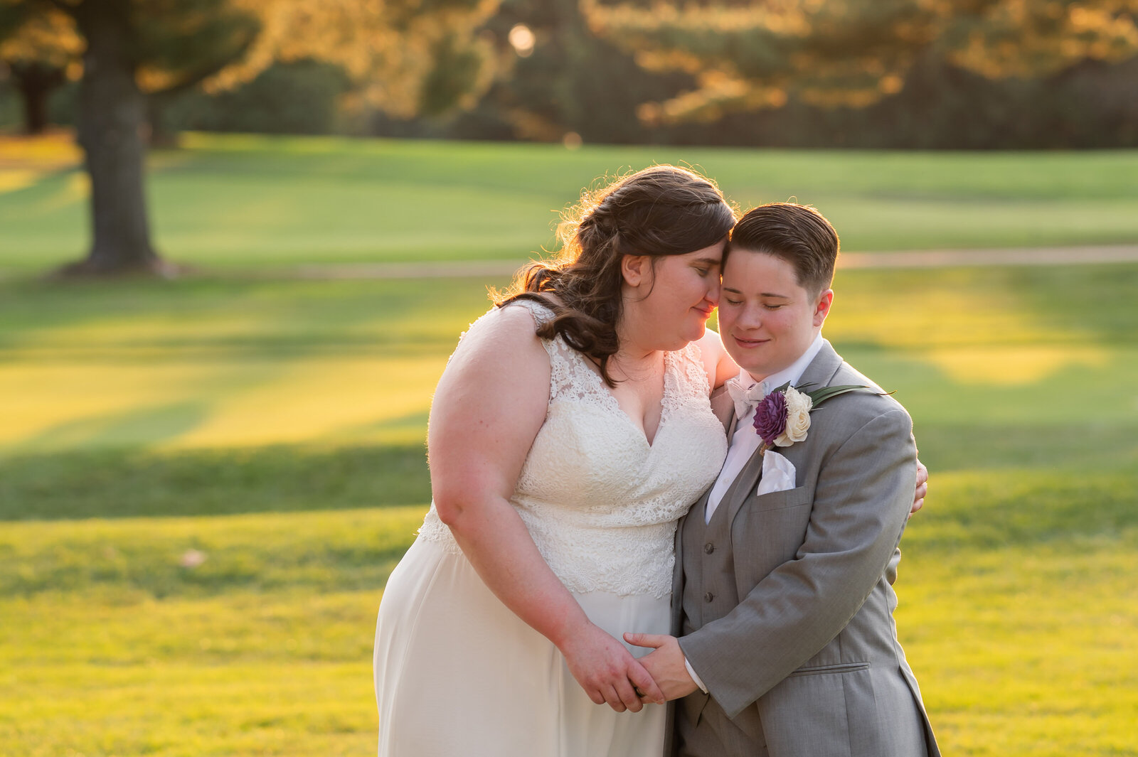 A couple in wedding attire stands close on a sunny lawn, with trees and sunlight in the background.