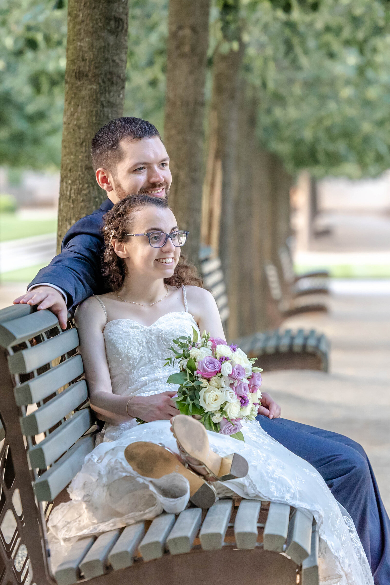 A bride and groom share a joyful moment sitting on a park bench surrounded by trees, with the bride holding a vibrant bouquet, captured during a Longwood Gardens, Pennsylvania wedding, highlighting authentic and romantic moments.