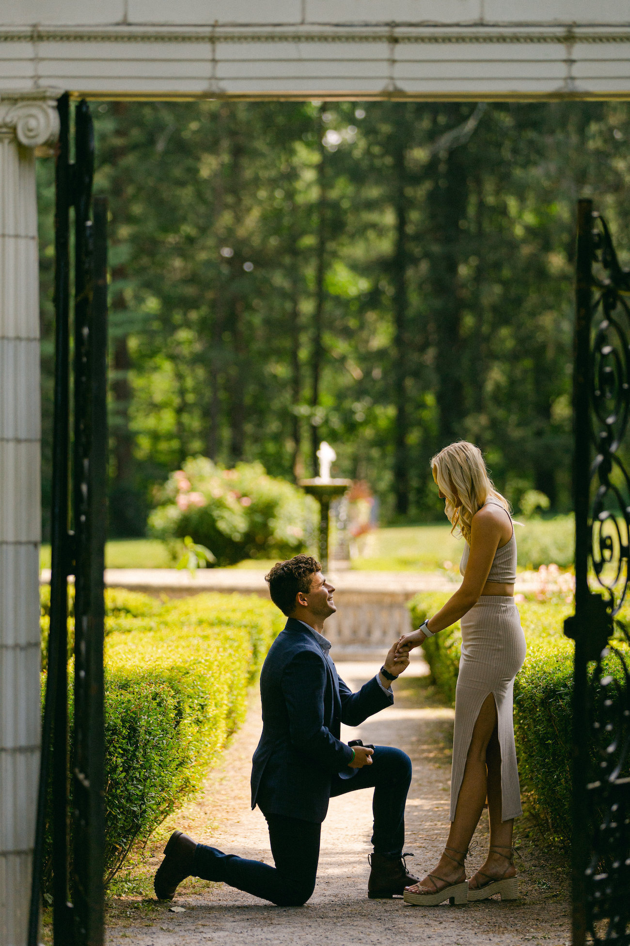A man proposing to a woman under a tall garden entrance gate with a fountain in the background.
