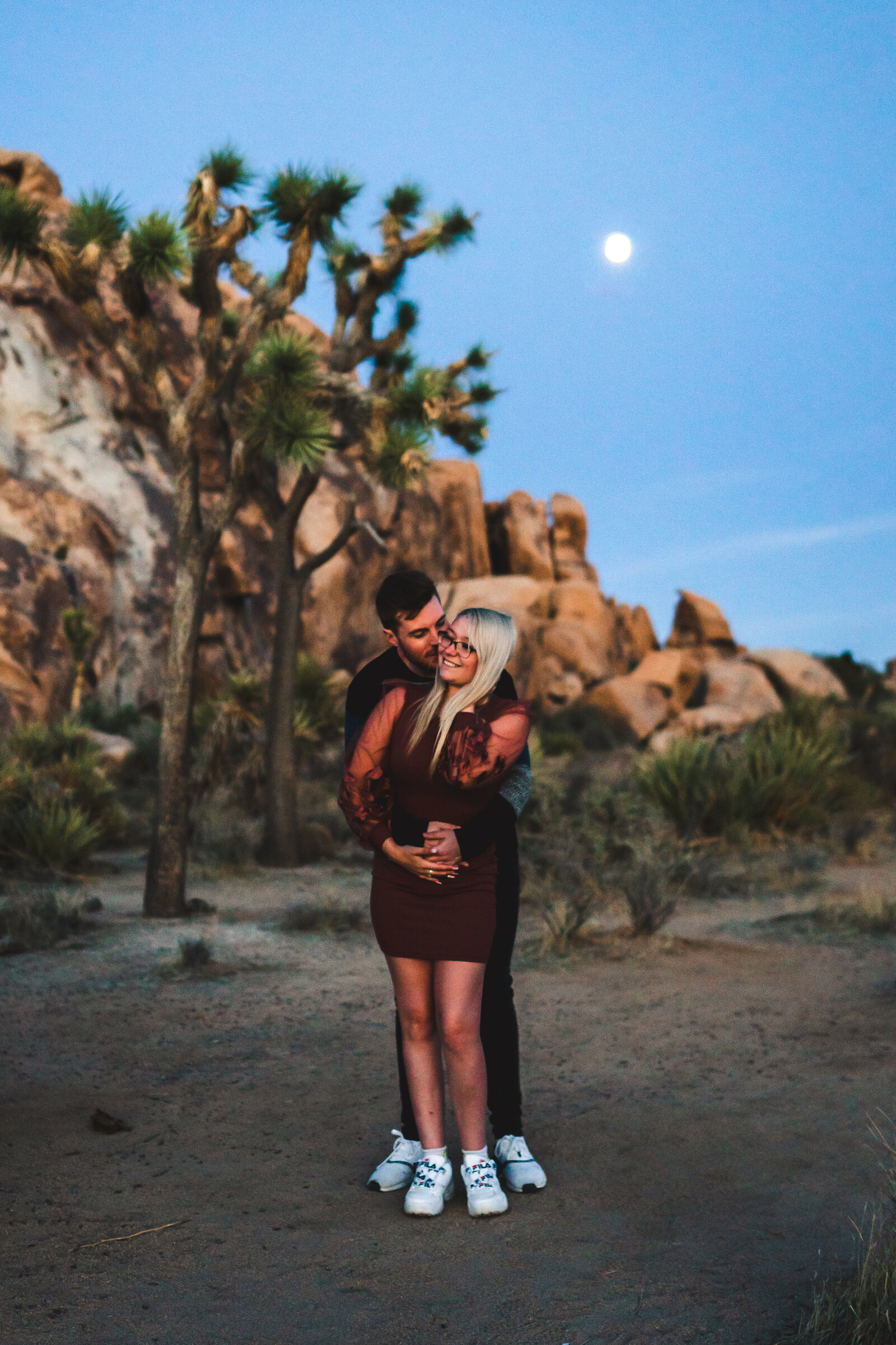 Couple holding each other, man kissing woman on cheek, with Joshua Trees behind them and moon rising.