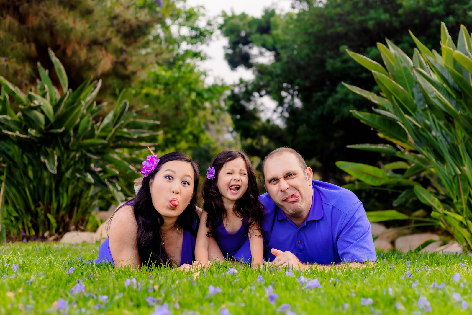 Family Photographer, a mother, father, and daughter make funny faces in the grass before flowers