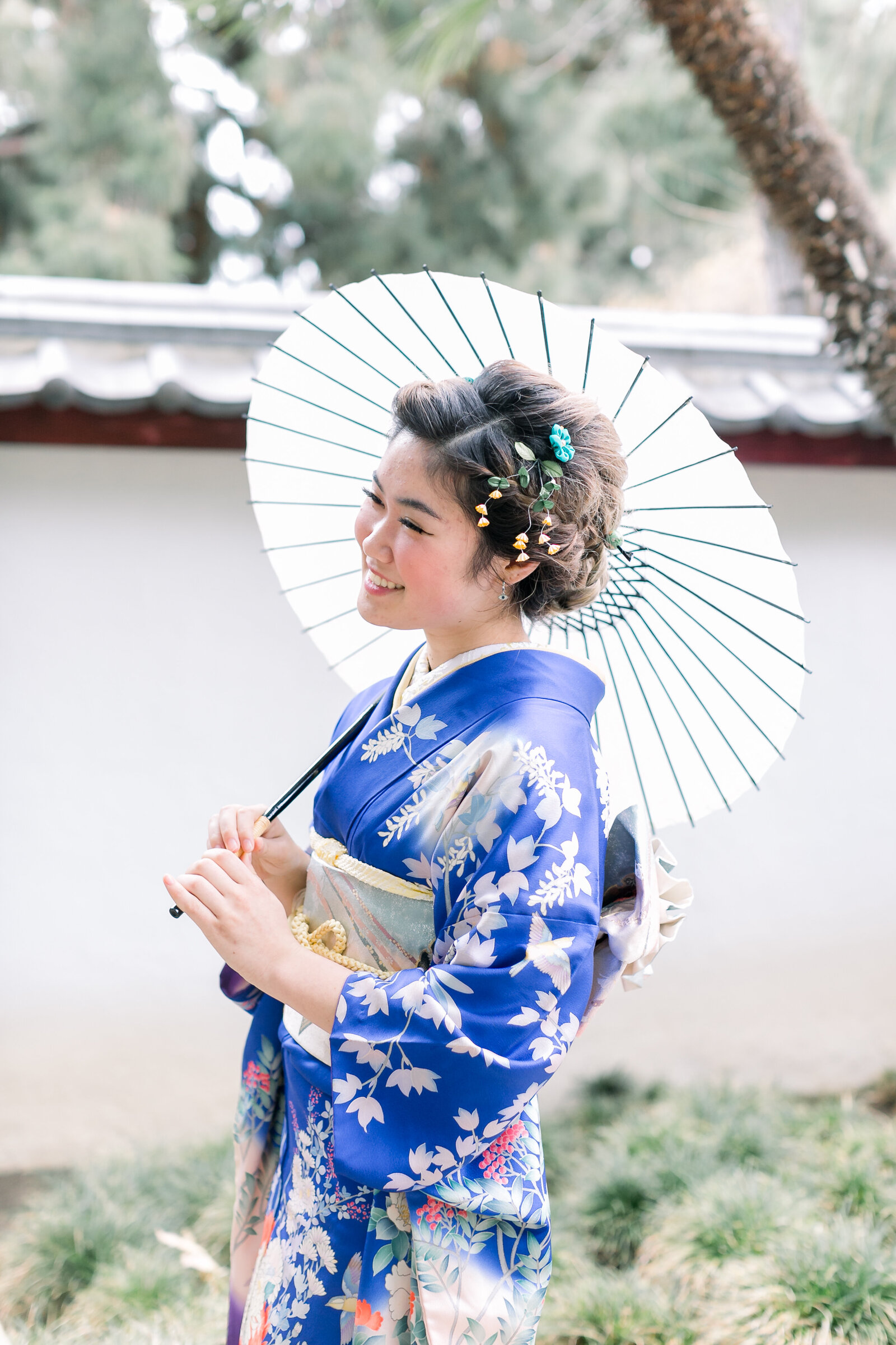 Smiling woman in blue floral furisode holding a traditional Japanese parasol during her Seijinshiki celebration at Brand Park Glendale. Available for kimono rental in Los Angeles | 成人式の青い振袖で和傘を手にブランドパーク（グレンデール）で微笑む女性、ロサンゼルスで着物レンタル