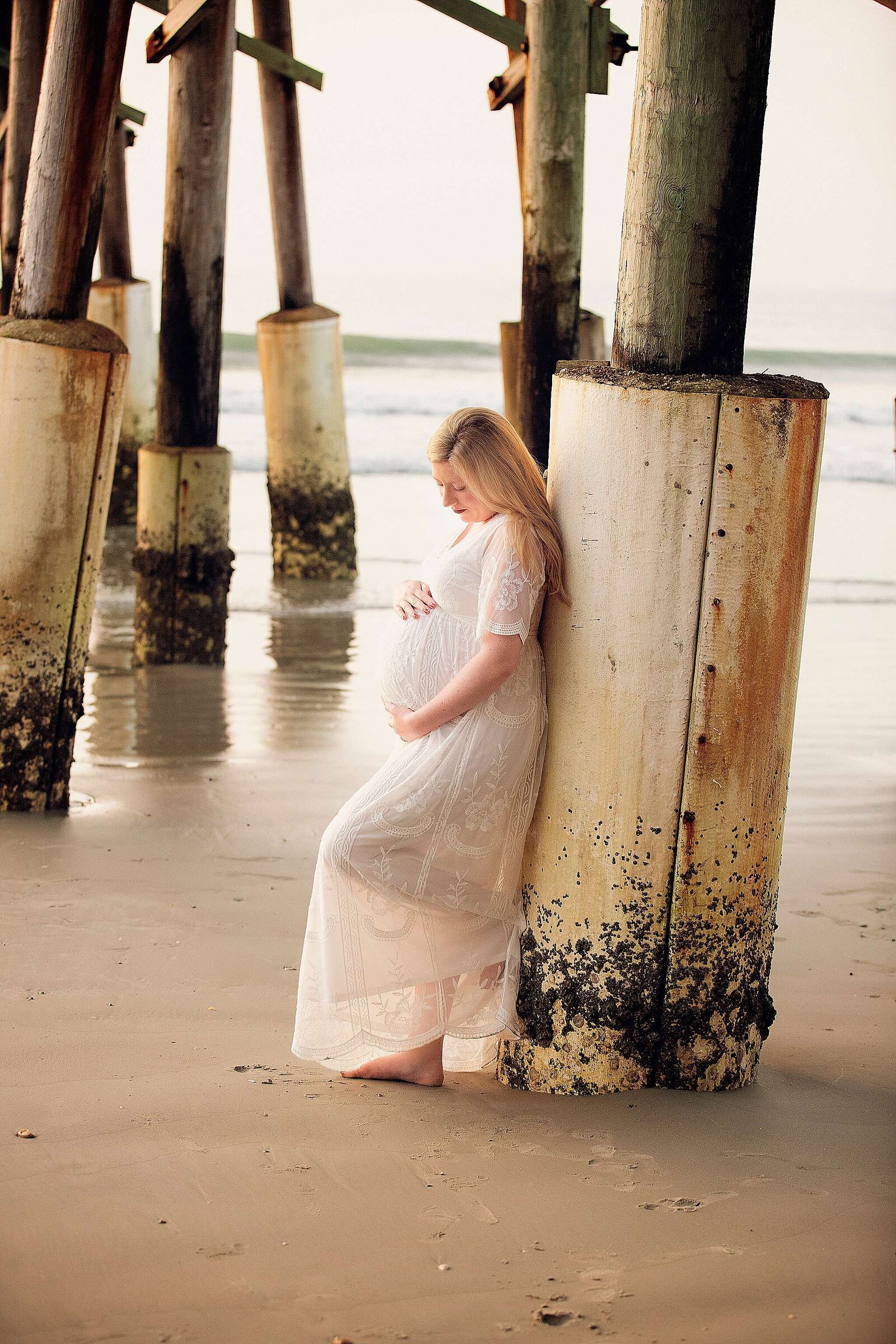 Sunrise maternity session at Cocoa Beach, FL, featuring a pregnant woman in a white dress leaning against a pillar.