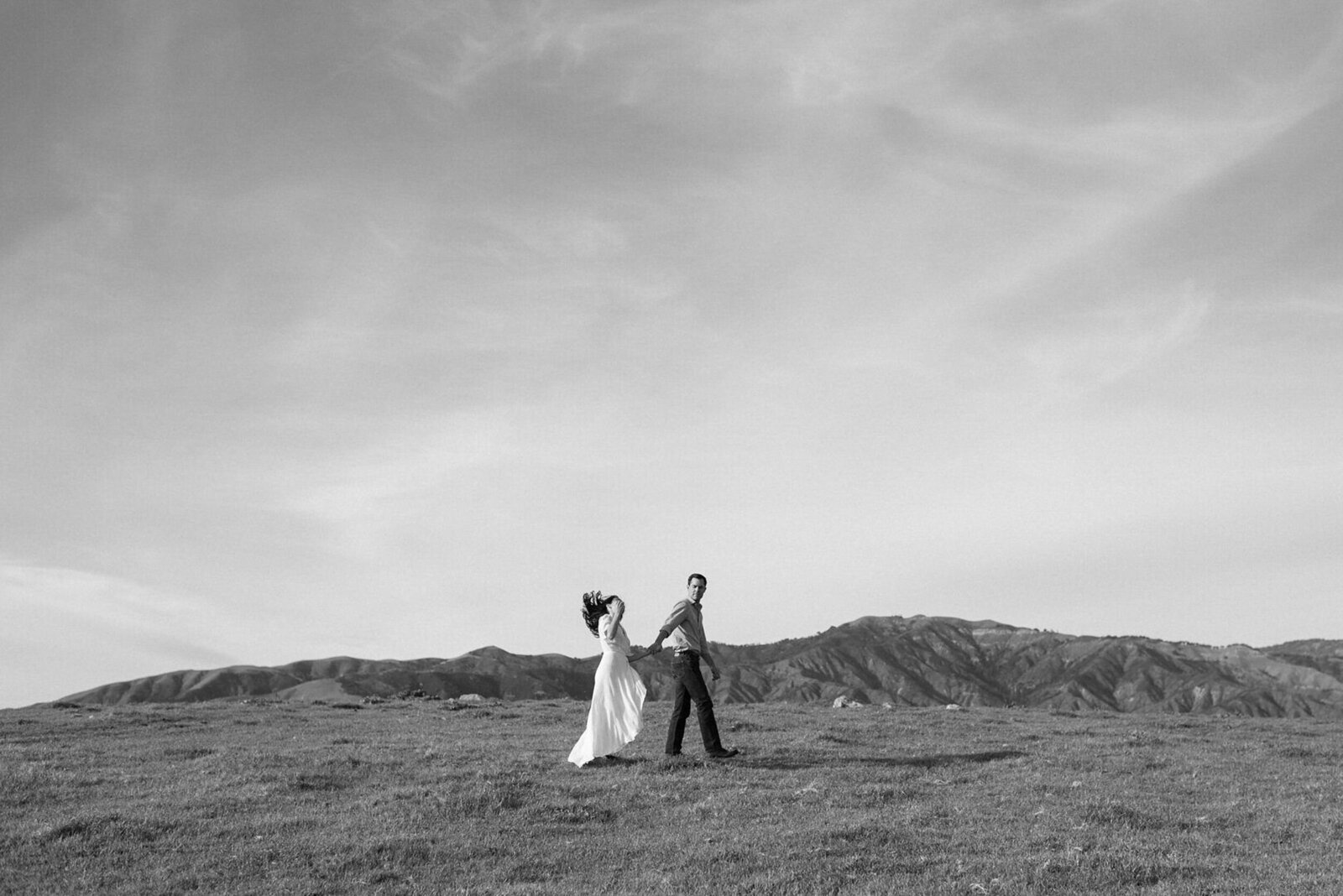 Quirky black & white portrait of couple small in frame walking through a Big Sur mountain scape with bride tossing her hair 