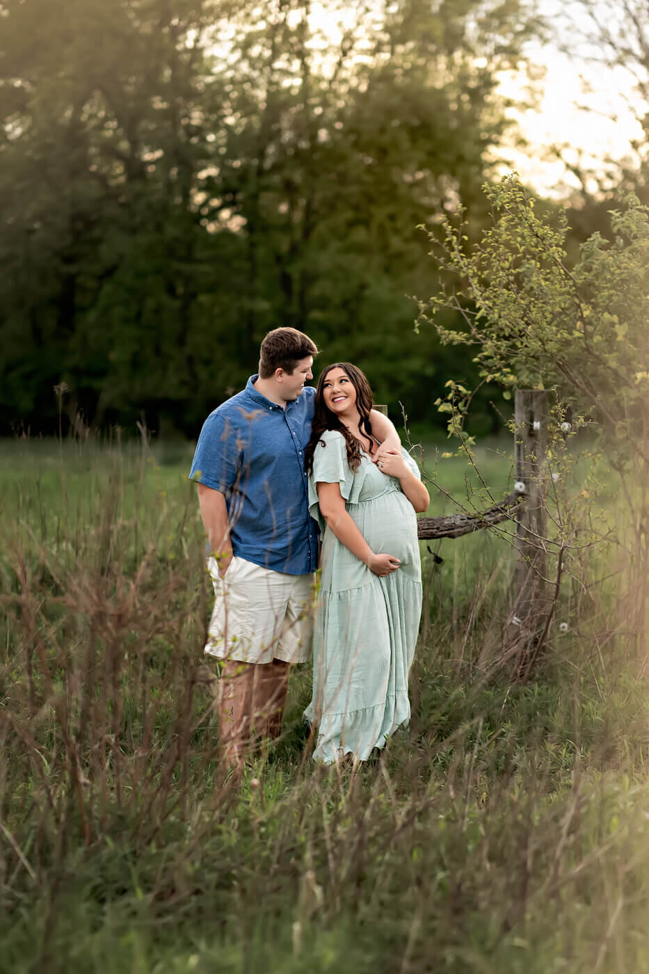 A smiling pregnant woman in a light green dress stands in a grassy field, embraced by her partner near a wooden fence and small tree, bathed in warm sunlight—capturing the joy of maternity portraits with a Bloomington maternity photographer.