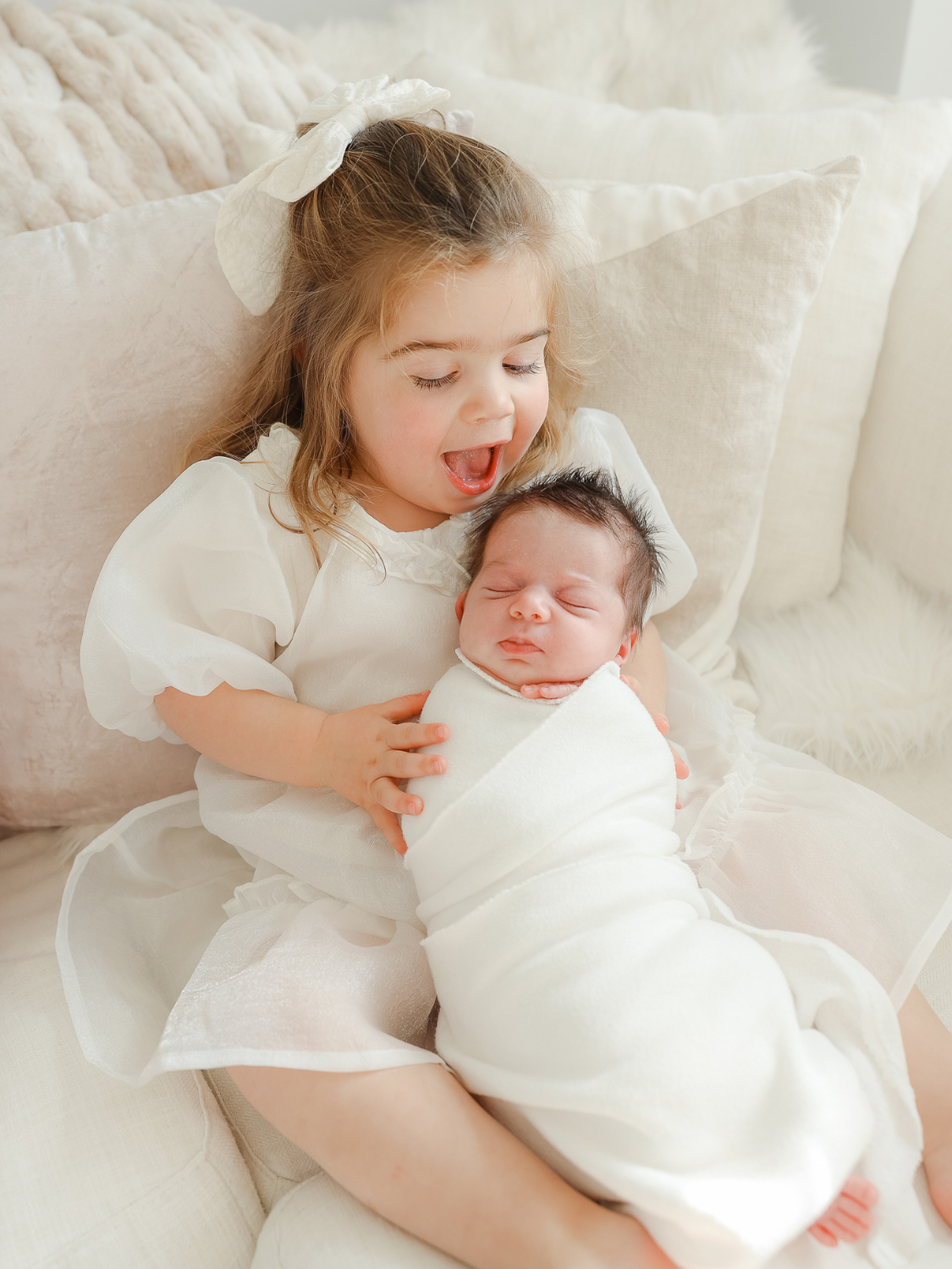 excited toddler wearing pretty white dress holds newborn sister during boston newborn photoshoot 