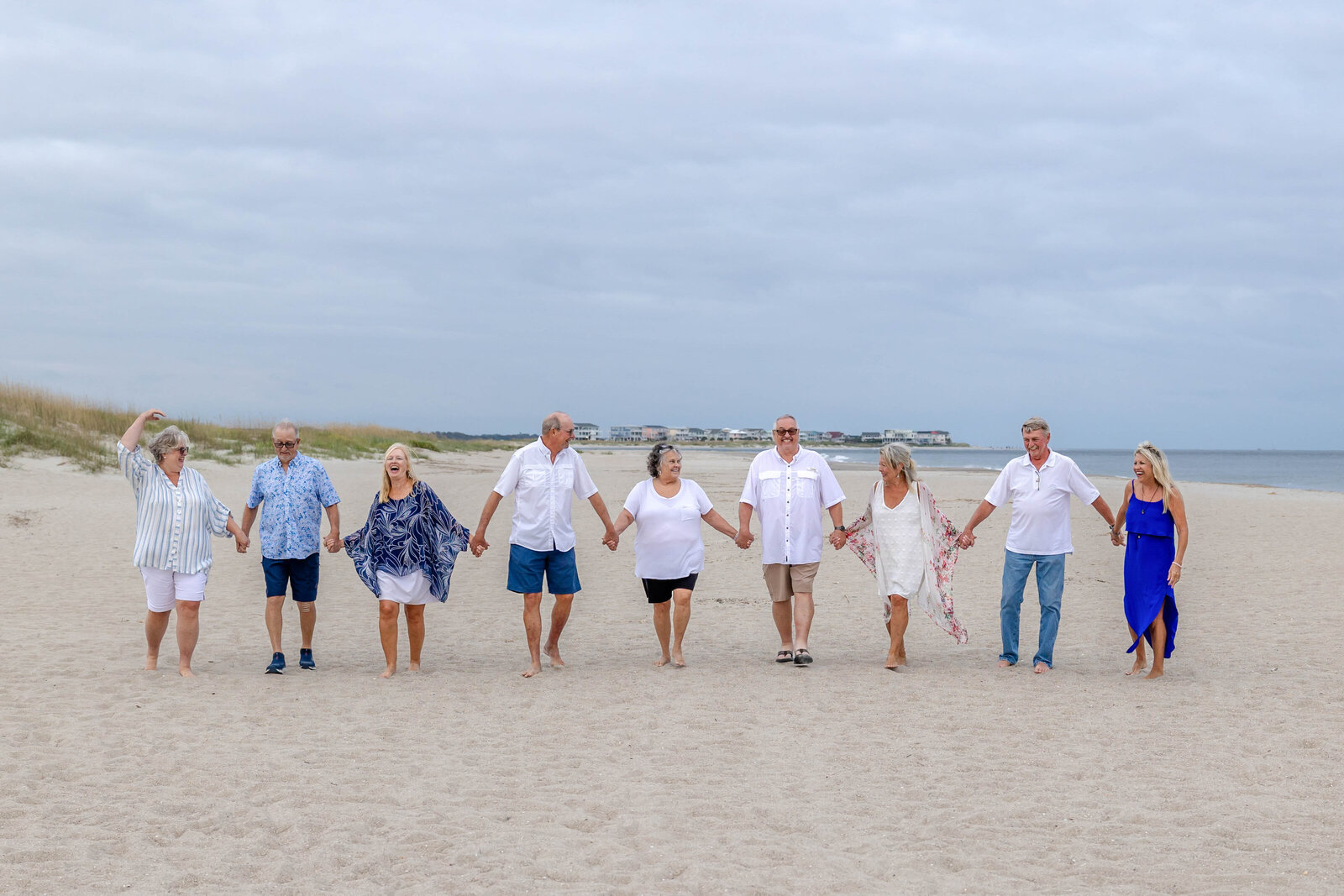 A group of adults walks hand in hand along a sandy beach, dressed in casual white and blue attire. The overcast sky adds a serene touch, ideal for professional family photography in NC, showcasing joyful connections and coastal charm.