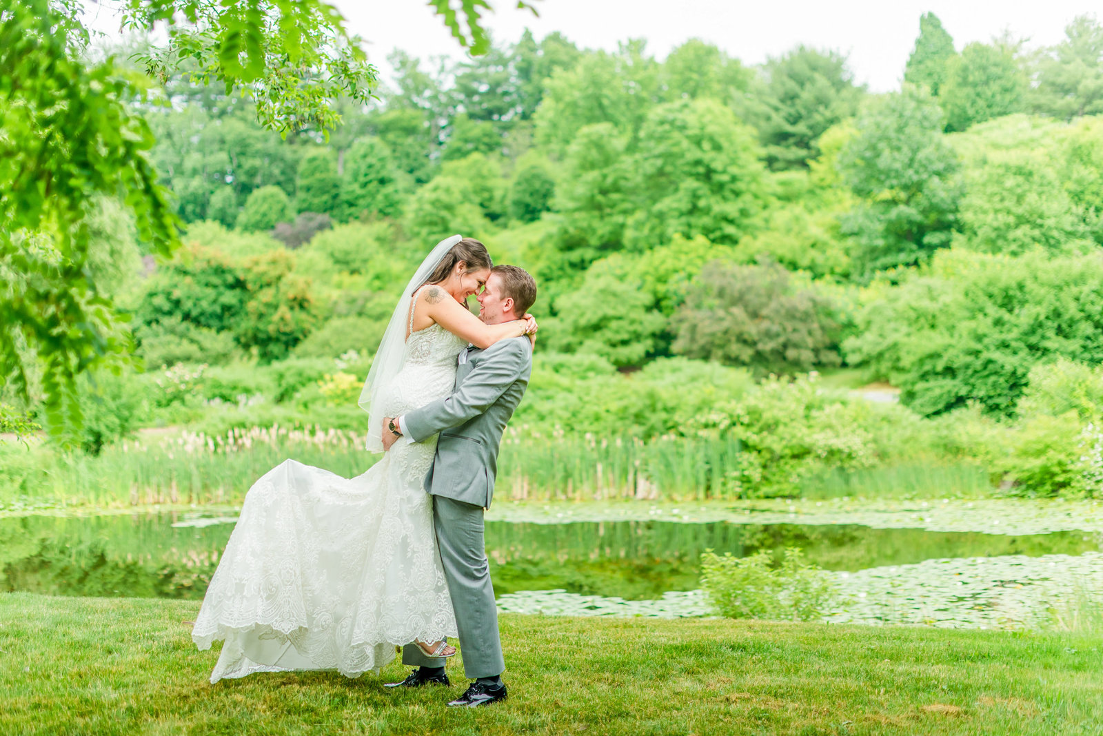 Groom lifts bride for a picture in front of a pond surrounded by green trees. Syracuse Photographer
