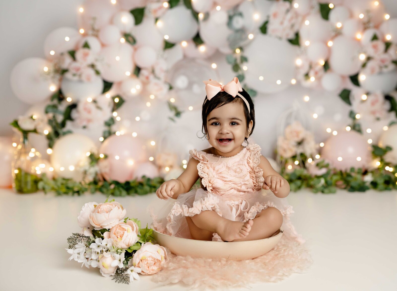 Baby girl sitting in a bowl in front of peachy pink balloon garland. 1st birthday photo taken by Jenni Nikki Photography in Atlanta, GA.