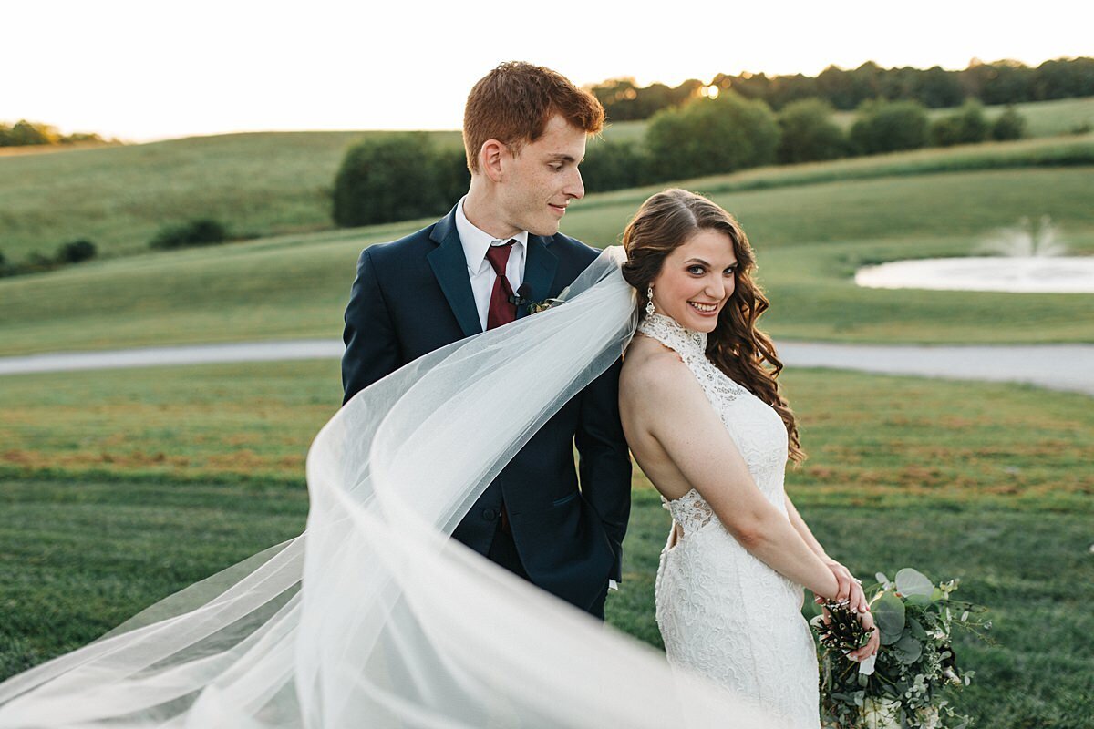 bride in a lace haltertop dress wearing a long veil blowing in the wind standing with her back turned to the groom who is wearing a dark blue suit and maroon tie is facing forward looking down at her smiling.