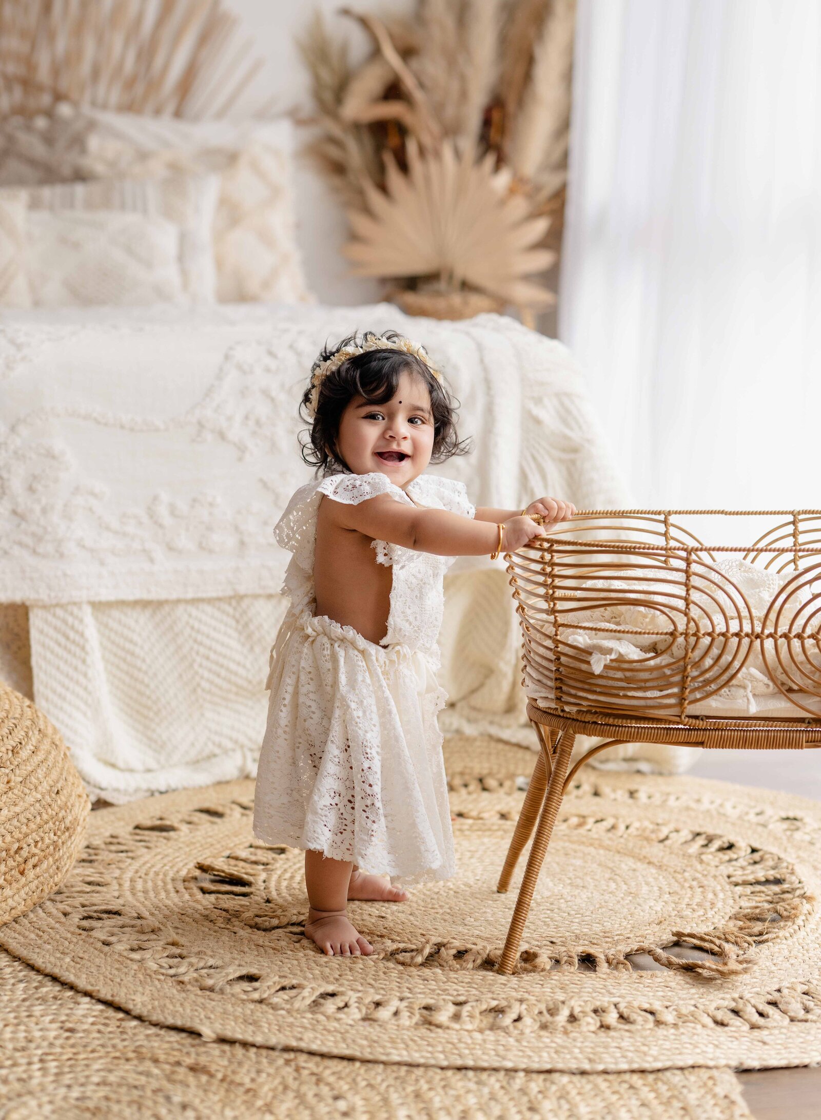 Baby girl standing up and holding onto bassinet that is in a boho lifestyle bedroom. Photographed at Atlanta photography studio.