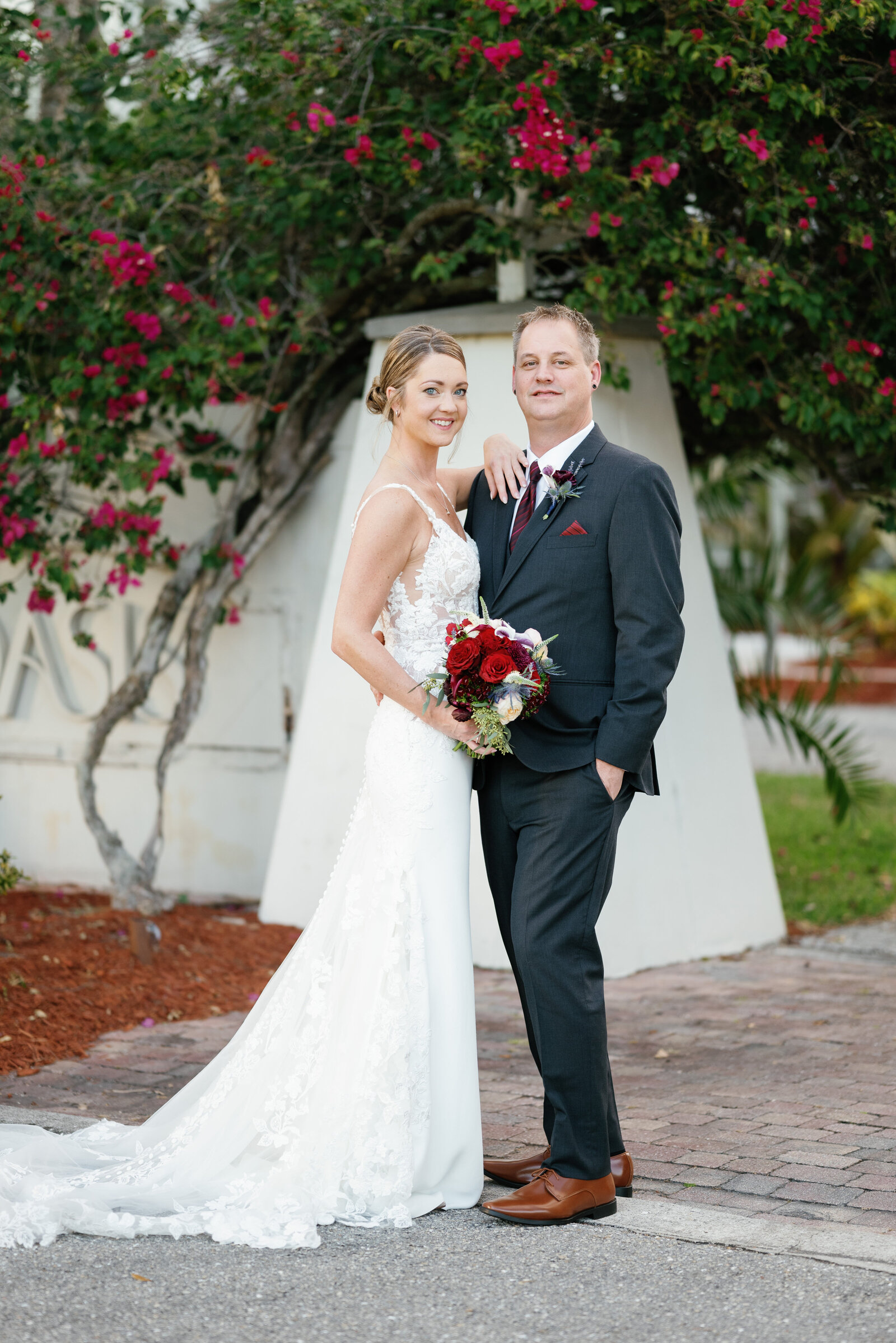 Bride and groom standing in front of a large bouganvilla bush, looking and the camera and smiling. Bride has her arm up on her grooms shoulder while his hand in in his pocket with his back arm wrapped around brides waist.