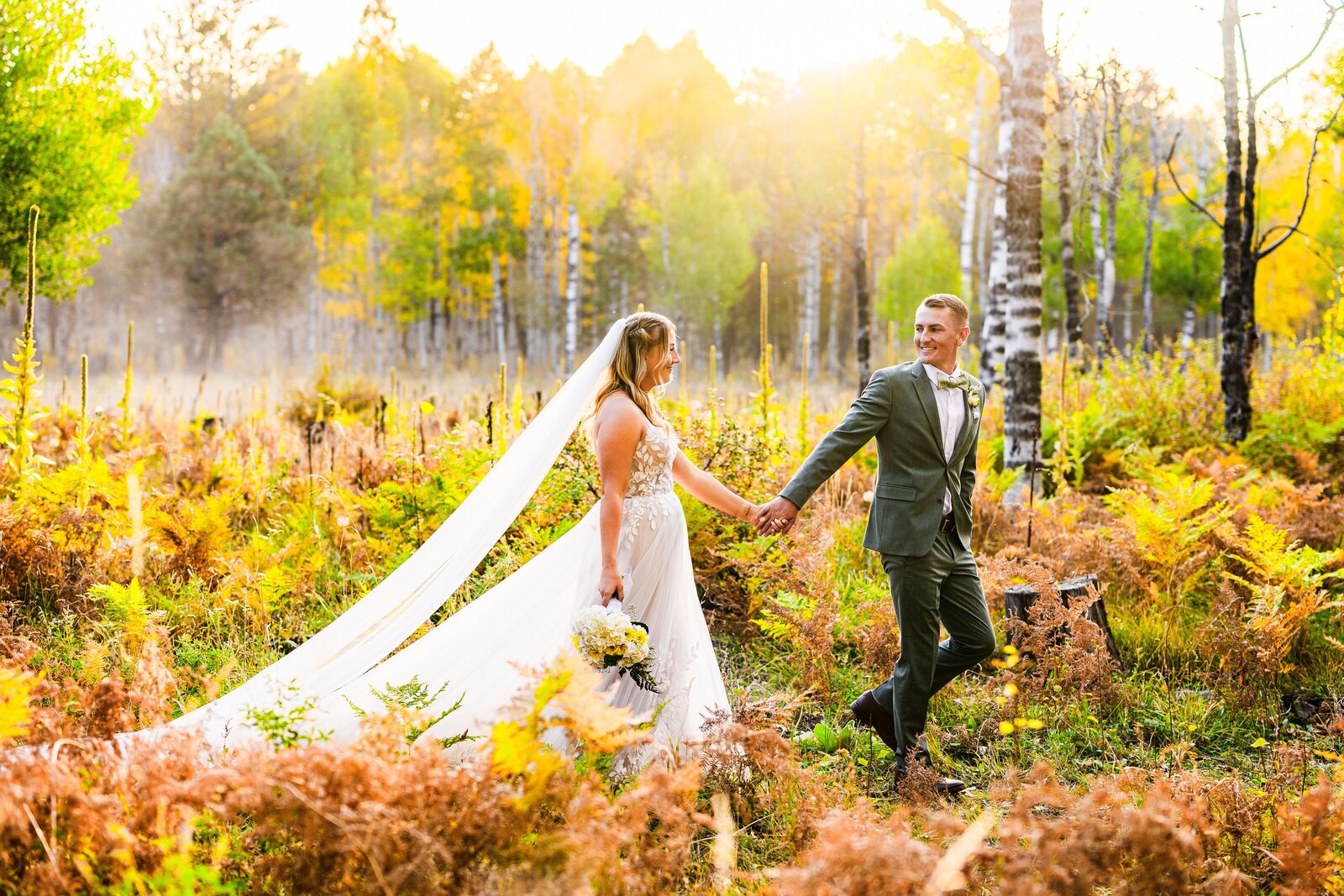 groom wearing green suit leading bride in wedding dress and long veil through aspen grove forest at golden hour in Flagstaff