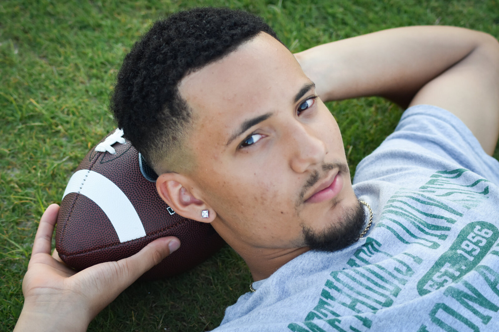 a senior posing for his graduation photos laying in the grass with a football under his head photographed by Millz Photography in Greenville, SC