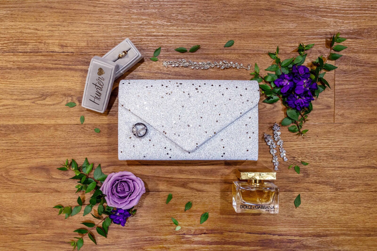 A bridal flat lay featuring a speckled clutch, Dolce & Gabbana perfume, a floral boutonniere, and wedding earrings, captured on wooden flooring during a Boone, NC wedding, showcasing elegant and creative details.
