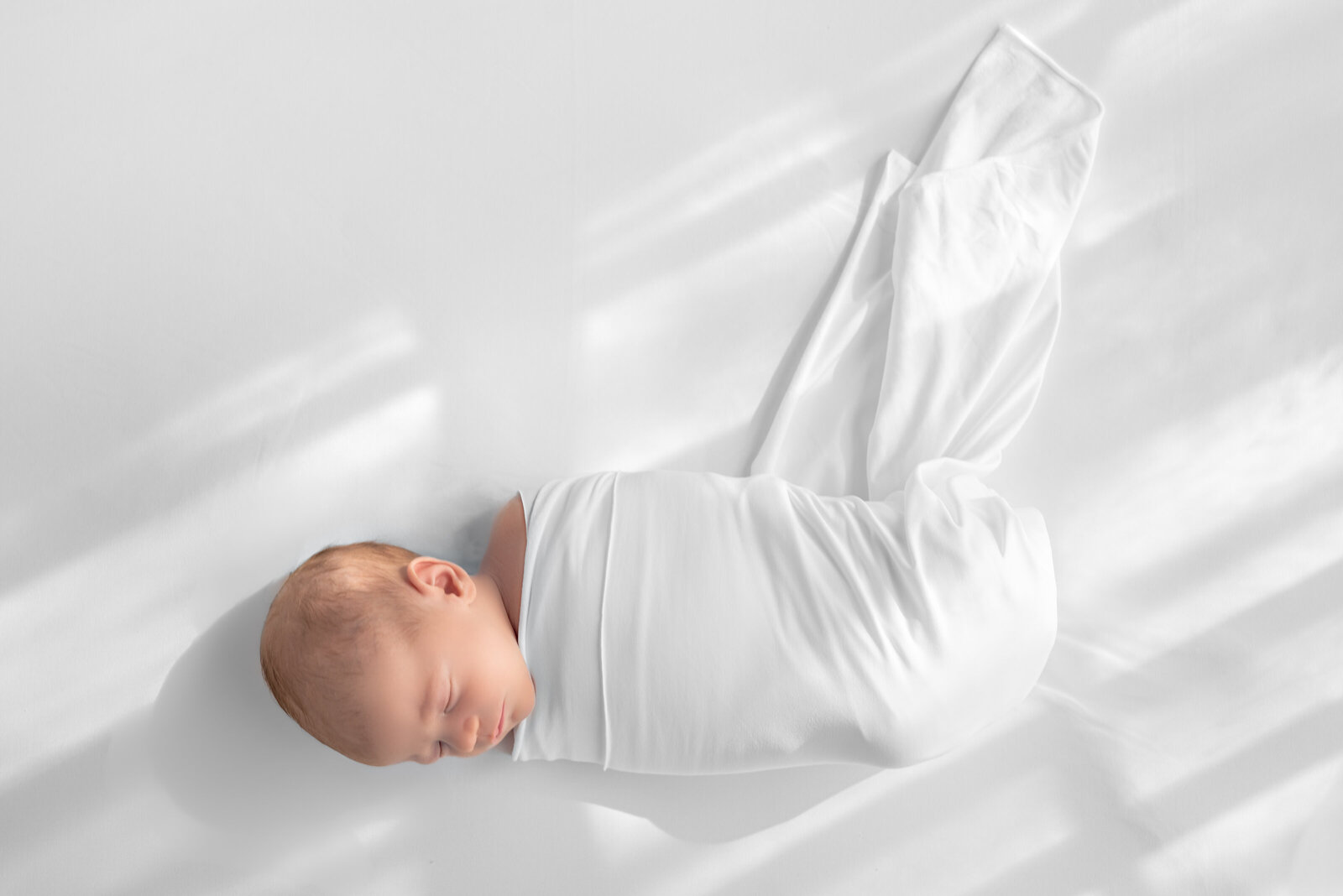 A newborn baby wrapped in a white blanket and sleeping for newborn photos in a studio in Hampton Cove  Alabama