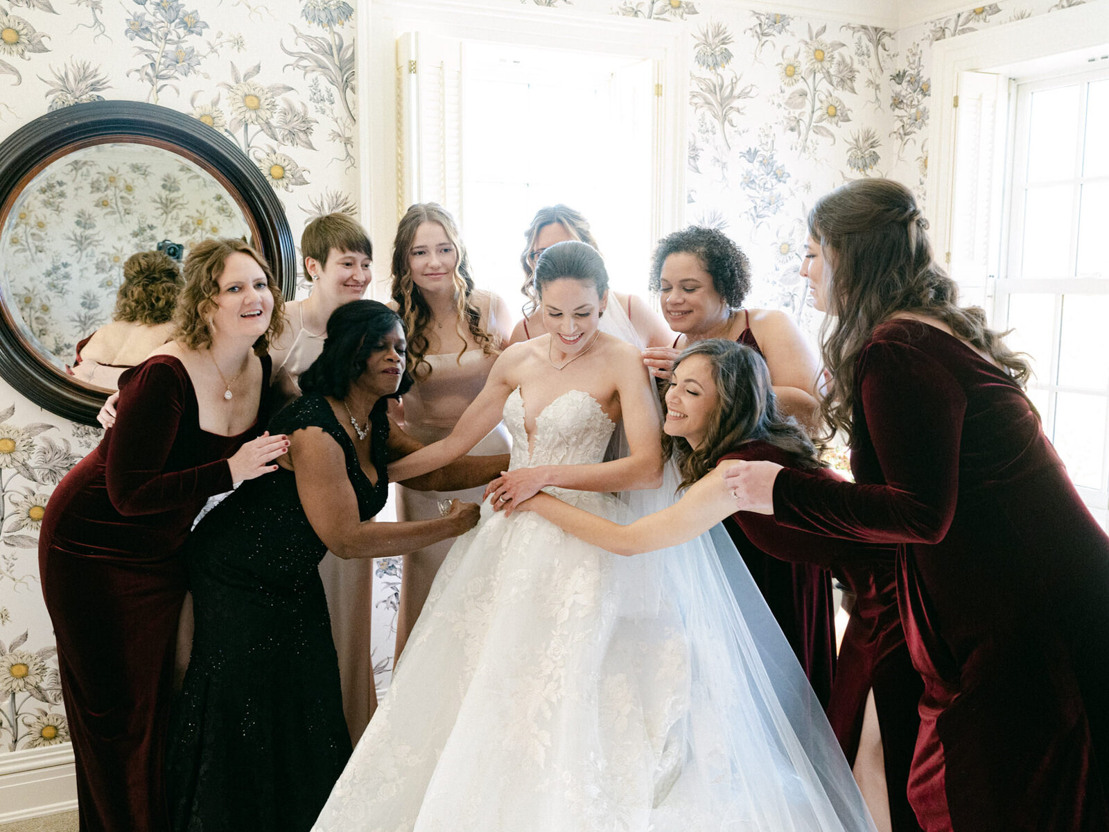 Bridesmaid in maroon velvet dresses hugging bride in white gown.