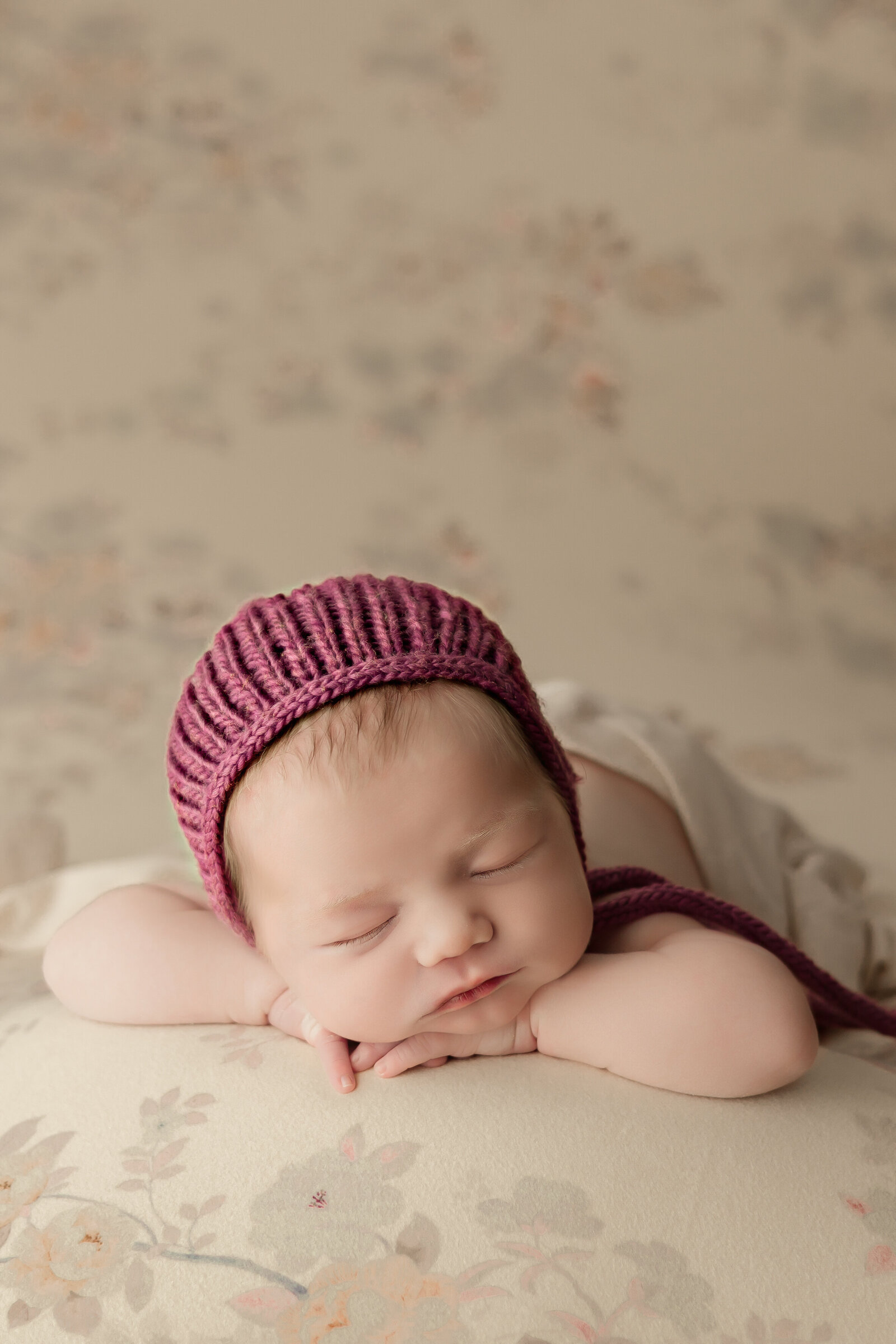 Newborn baby girl in plum knit bonnet on a soft floral background sleeping in her Harrisburg studio newborn photos