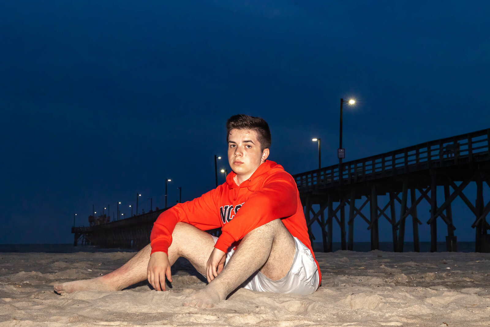 A high school senior wearing a red hoodie sitting on the beach at night with a pier illuminated in the background in North Topsail Beach, North Carolina. This moody portrait captures the unique atmosphere of senior photography, perfect for evening beach sessions.