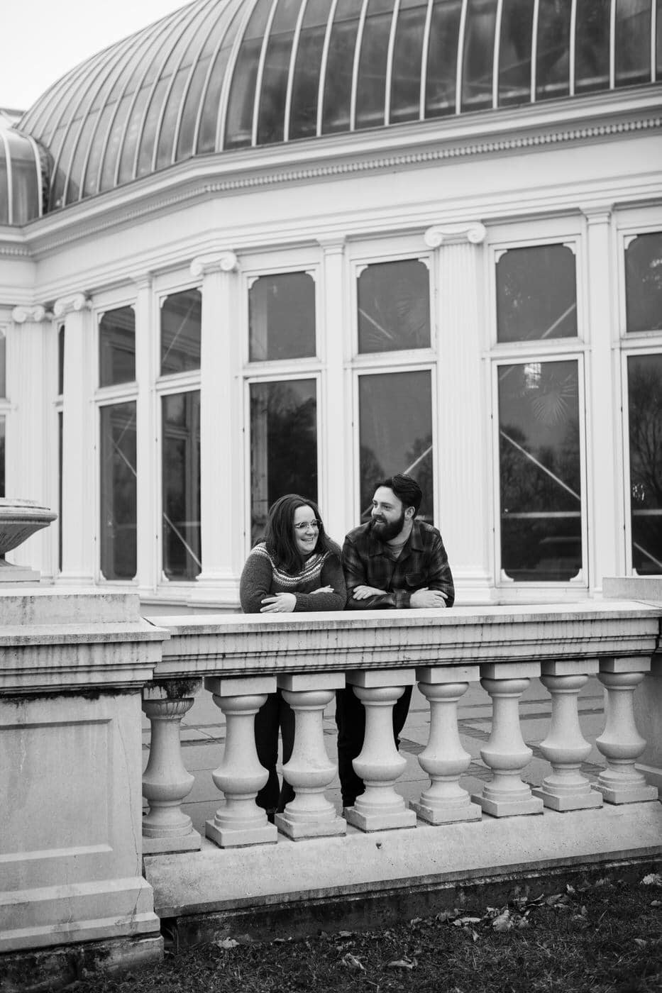 A couple leans side by side against a stone railing in front of a conservatory.