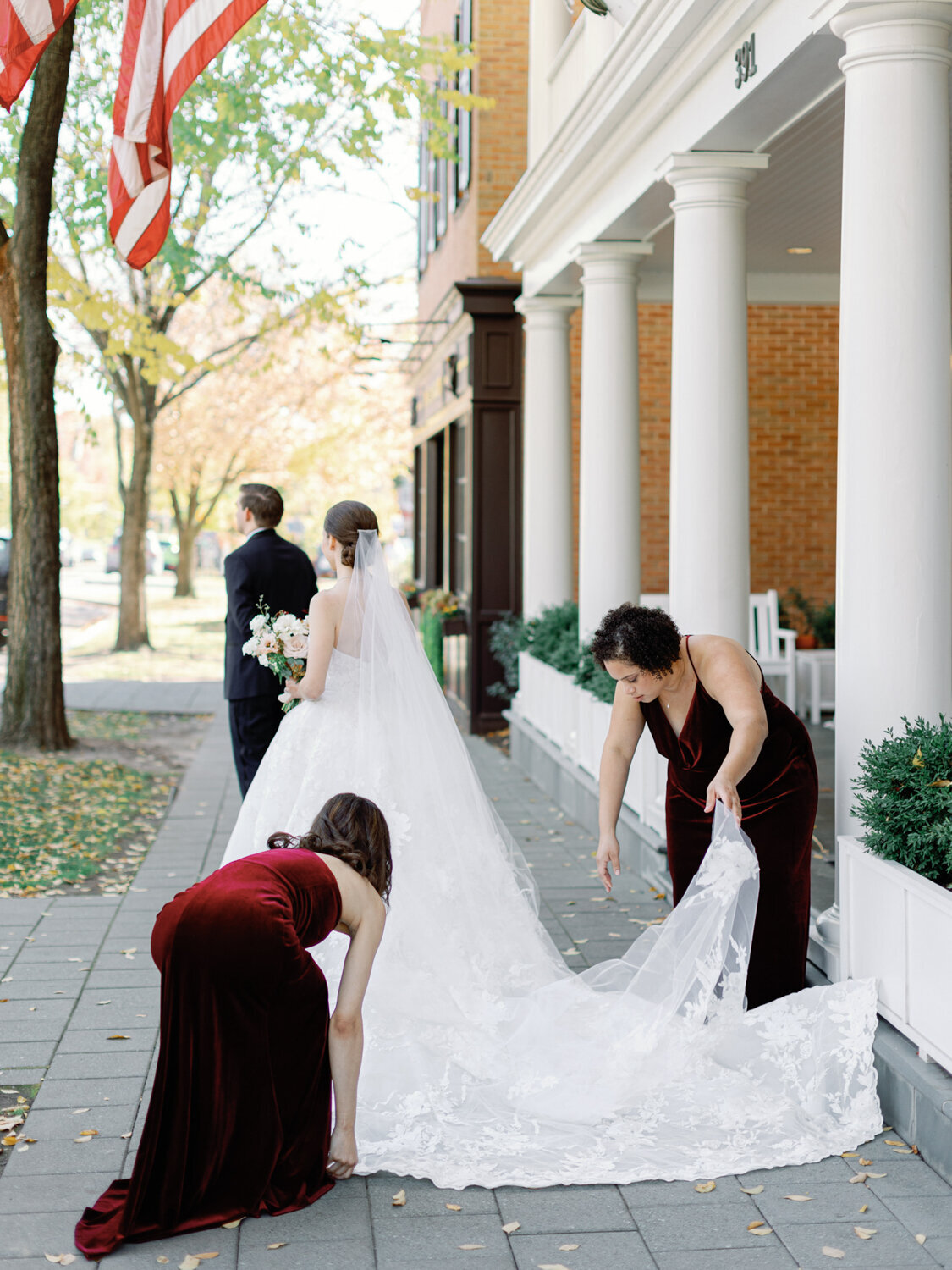 Bridesmaids help bride with her dress train as groom waits to see her for the first time.