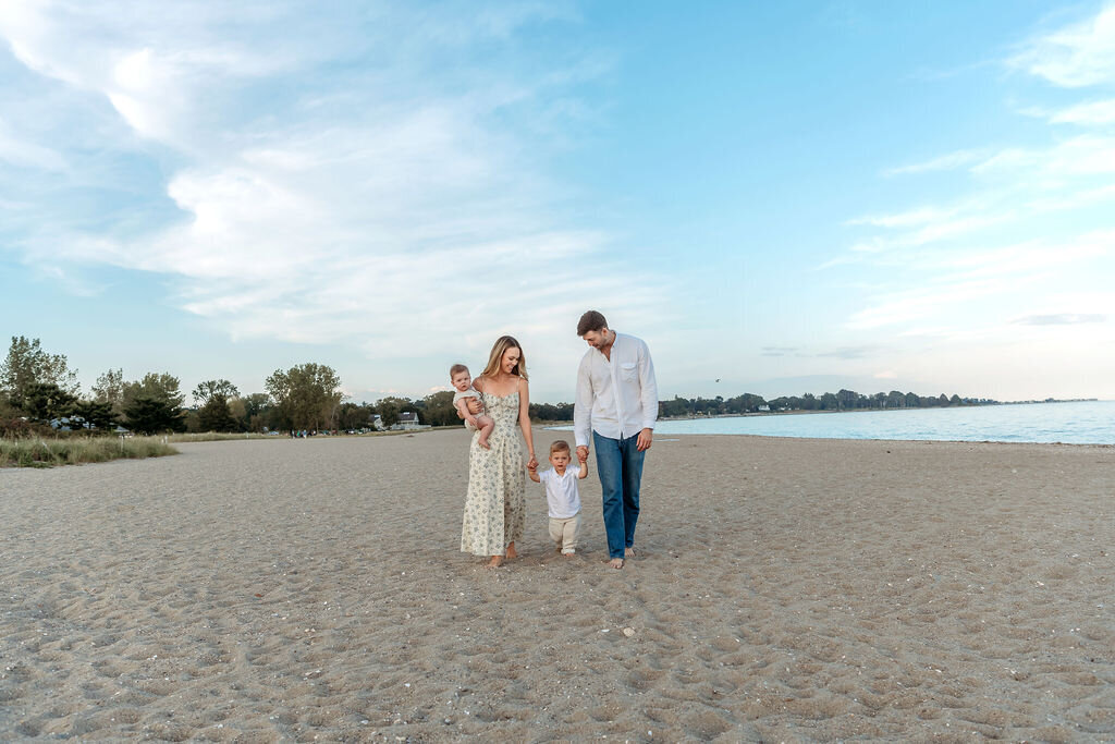 Connecticut Family Photographer captures a family of four walking hand-in-hand on a sandy beach at golden hour, with parents holding their toddler and young son against a backdrop of calm waters and cloudy skies