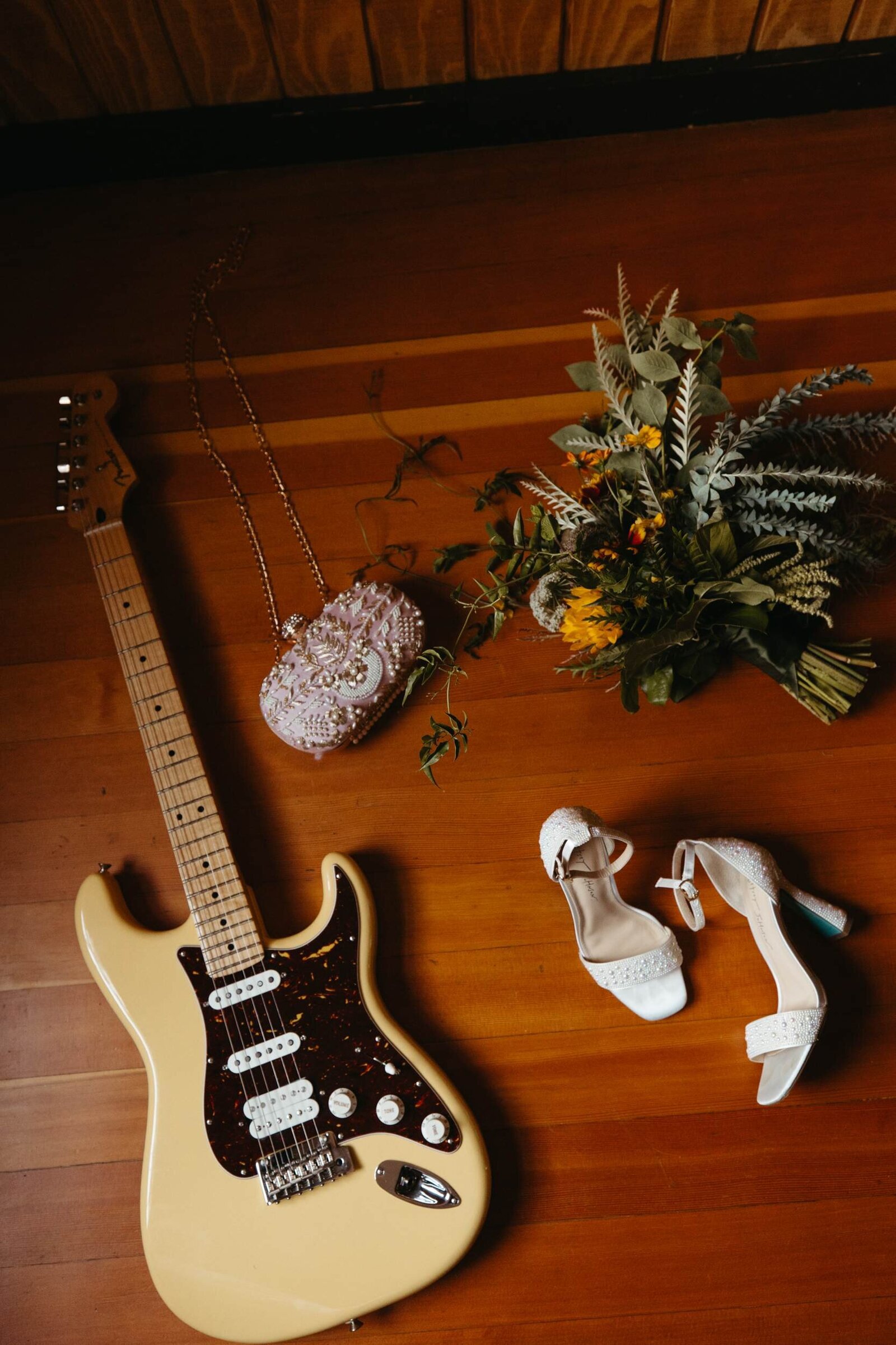 wedding details scattered artfully on wood floor including a guitar, white heels, beaded purse, and wild floral bouquet