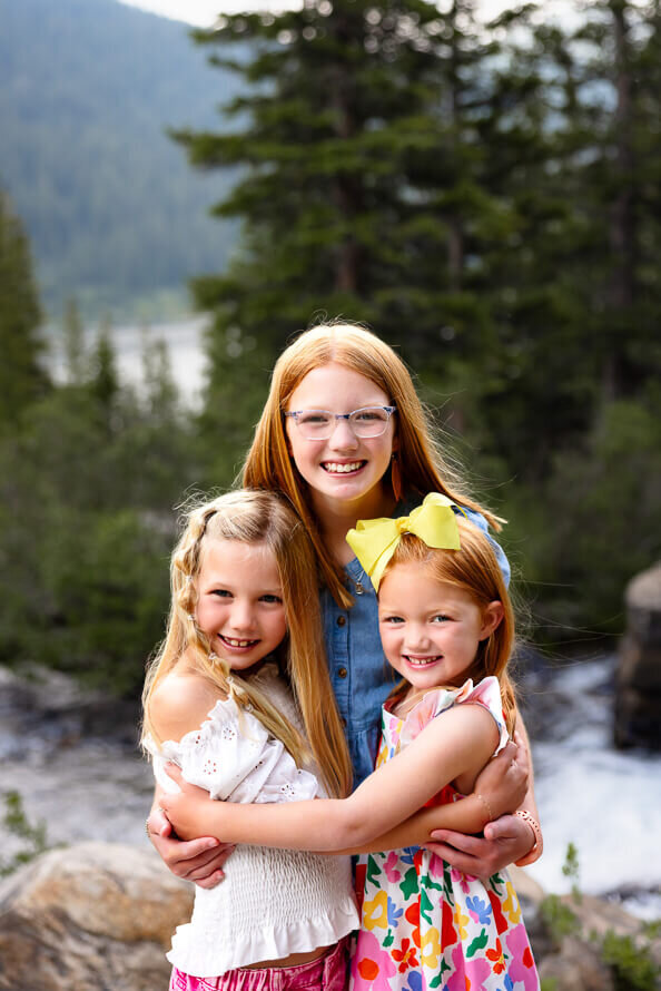 Three sisters hug and smile with trees and river and a lake in the background.
