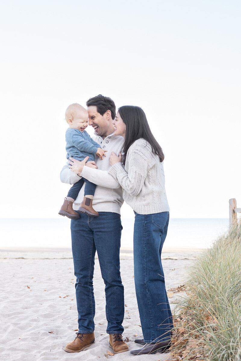 Young family with a baby smiling and playing on the beach during a lifestyle family photo session in Westchester County. Captured by Nicole, a CT family photographer who specializes in natural, heartfelt moments by the coast.
