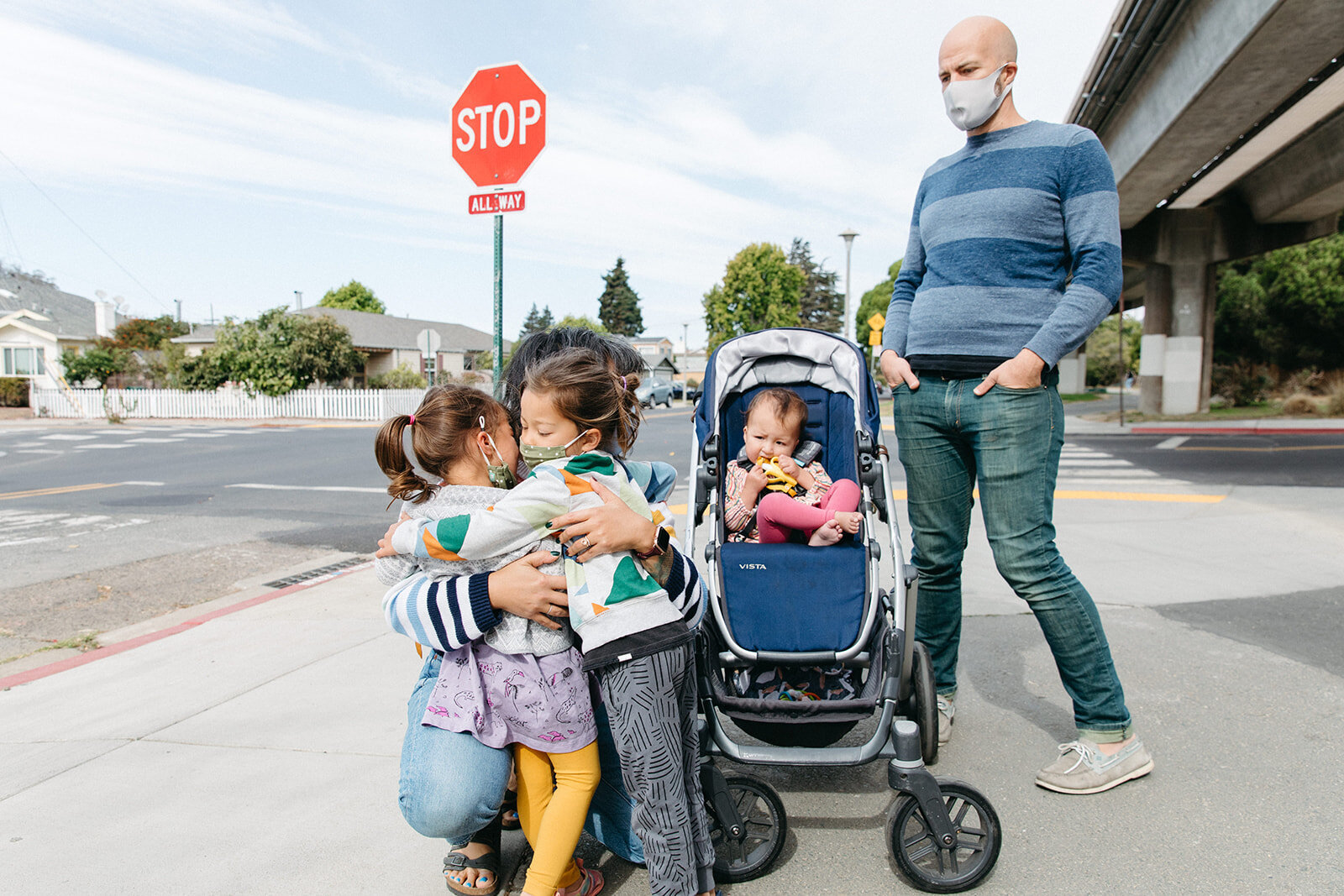 family of five standing on a corner in front of a stop sign with mom hugging two young kids with the third in a stroller while dad looks on