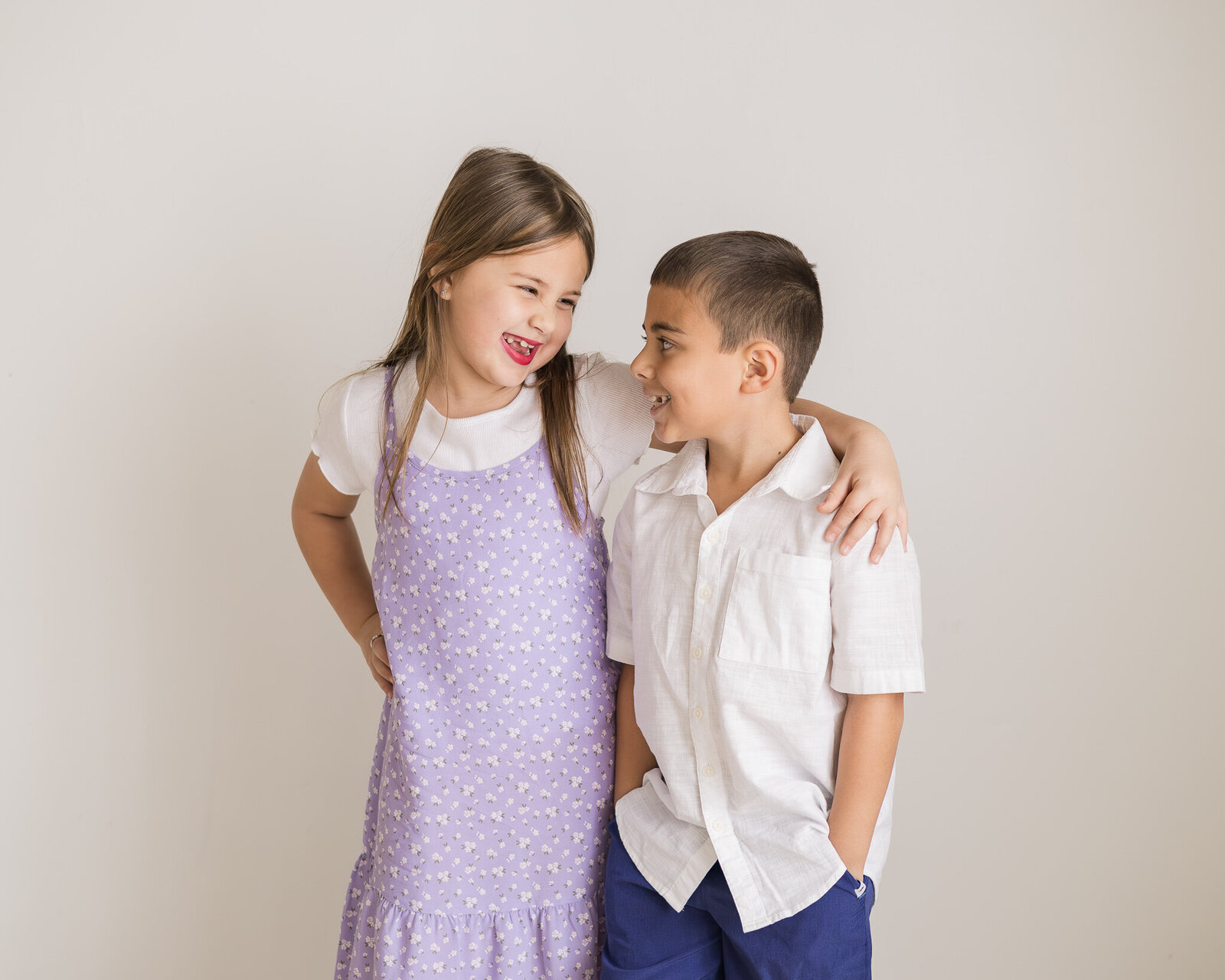 Brother and sister standing in a studio room, looking at each other and smiling.  Sister is wearing a light purple dress with a white shirt under it with dark brown hair  Brother is wearing a white button up shirt with blue shorts and dark brown hair.