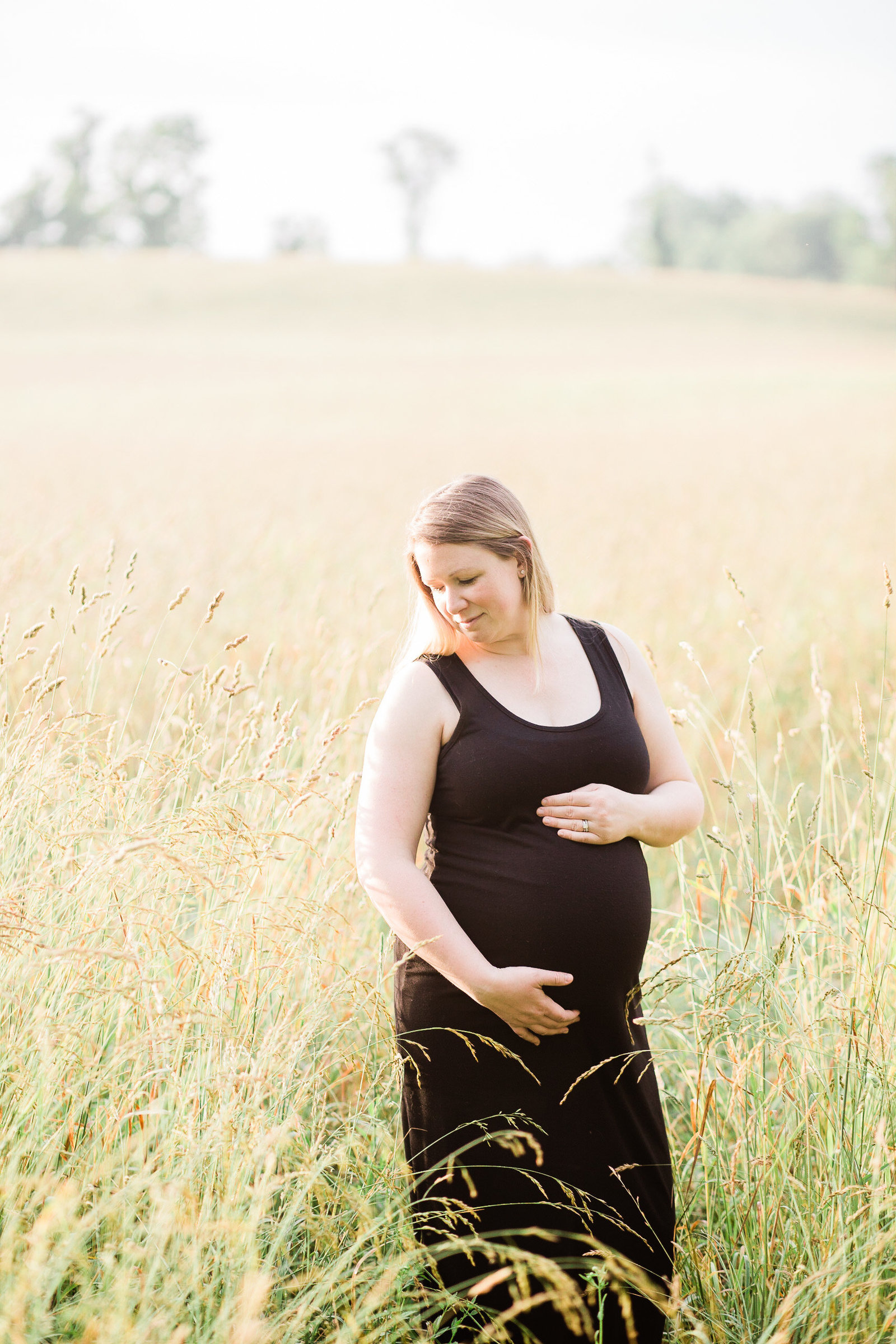 Blonde pregnant mother standing in a golden field in a black maxi dress, holding her baby bump