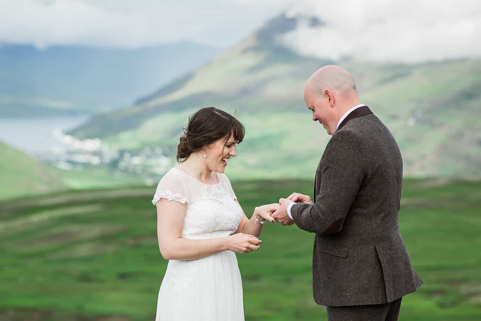 A groom slips the wedding band on a bride's finger during their ceremony overlooking a lush, green valley of Iceland. 
