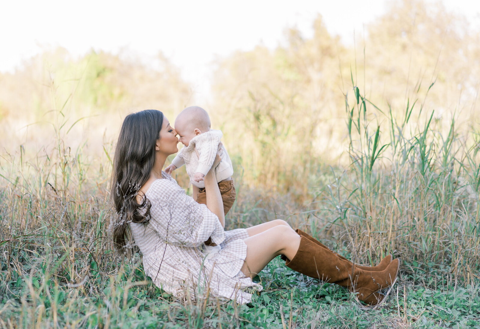 Portrait of a woman in a white dress and brown boots sitting in a country field holding and kissing a baby boy.