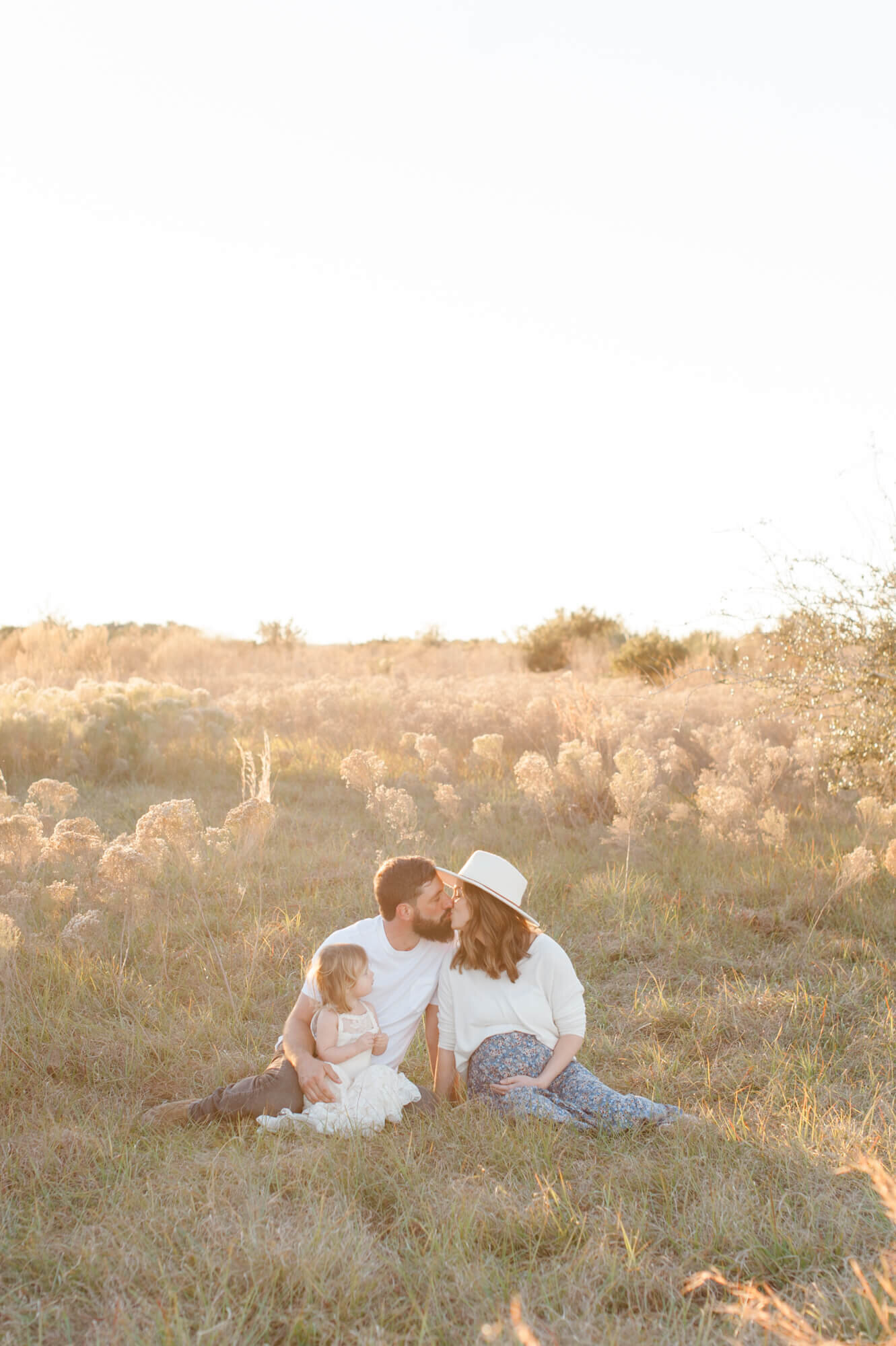 Pregnant couple kissing in a tall grass field while holding their young daughter during golden hour