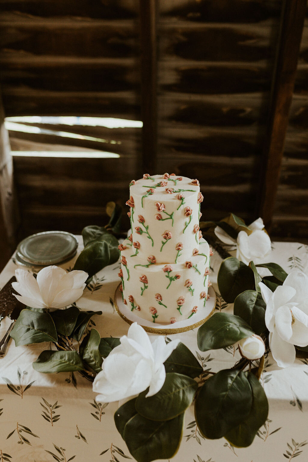 White wedding cake with pink and green frosting flowers sitting on a table surrounded by white flowers and greenery