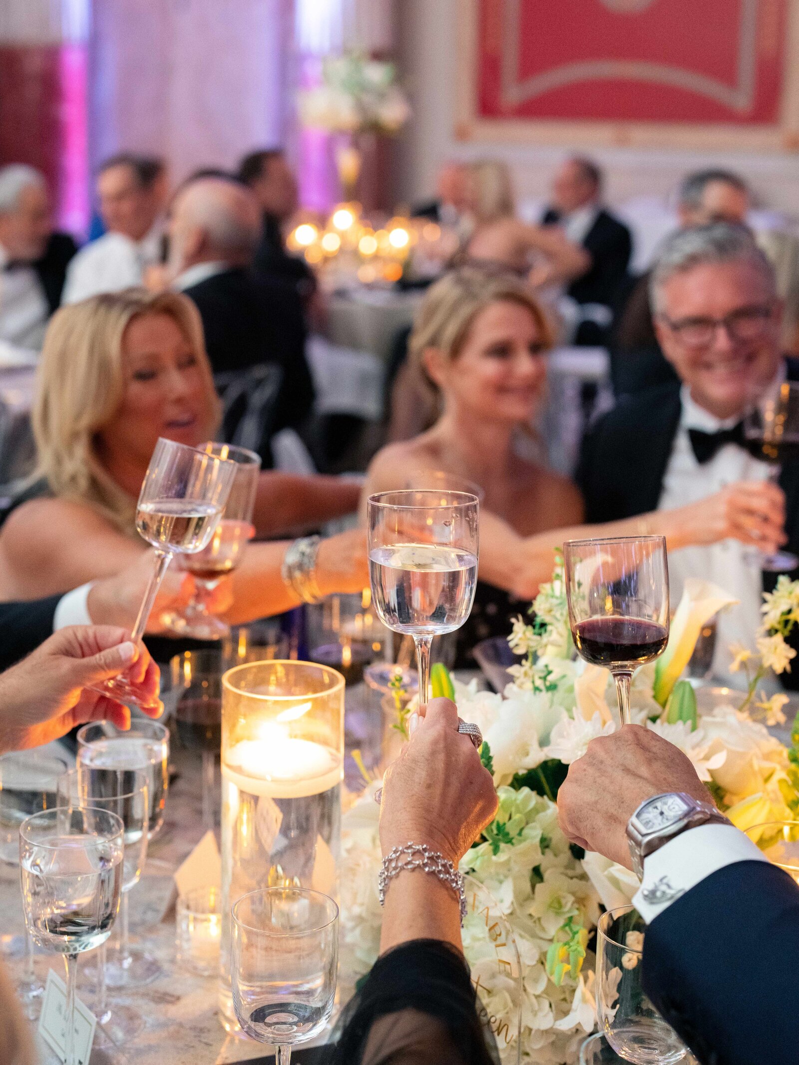 A group of people dressed elegantly in formal attire celebrate at a banquet table, raising glasses in a toast. The table is adorned with white flowers, candles, and glassware, creating a festive and elegant atmosphere.
