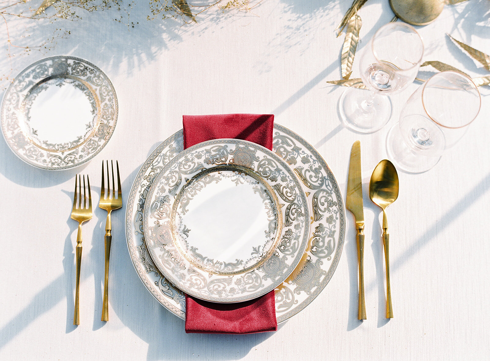 Table setting at luxury wedding reception. Ivory table linens with ivory dinner, salad and bread plate with intricate gold detailing. Brushed gold flatware, clear glasses with gold trim. Deep red velvet napkin wrapped around dinner plate with salad plate on top. Photographed at Lowndes Grove wedding reception in full sun with the shadows falling on the linens from the candles, photographed by wedding photographers in Charleston Amy Mulder Photography.