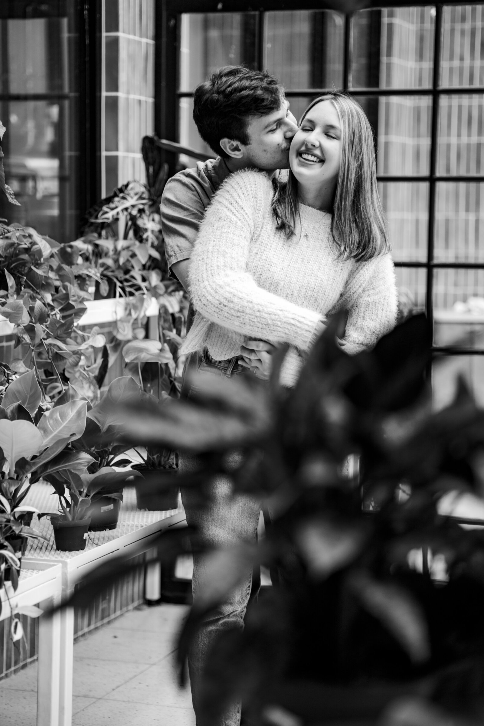 Black and white engagement session with Megan and Luke in a greenhouse next to Comeback Coffee in Memphis, embracing behind lush green leaves.