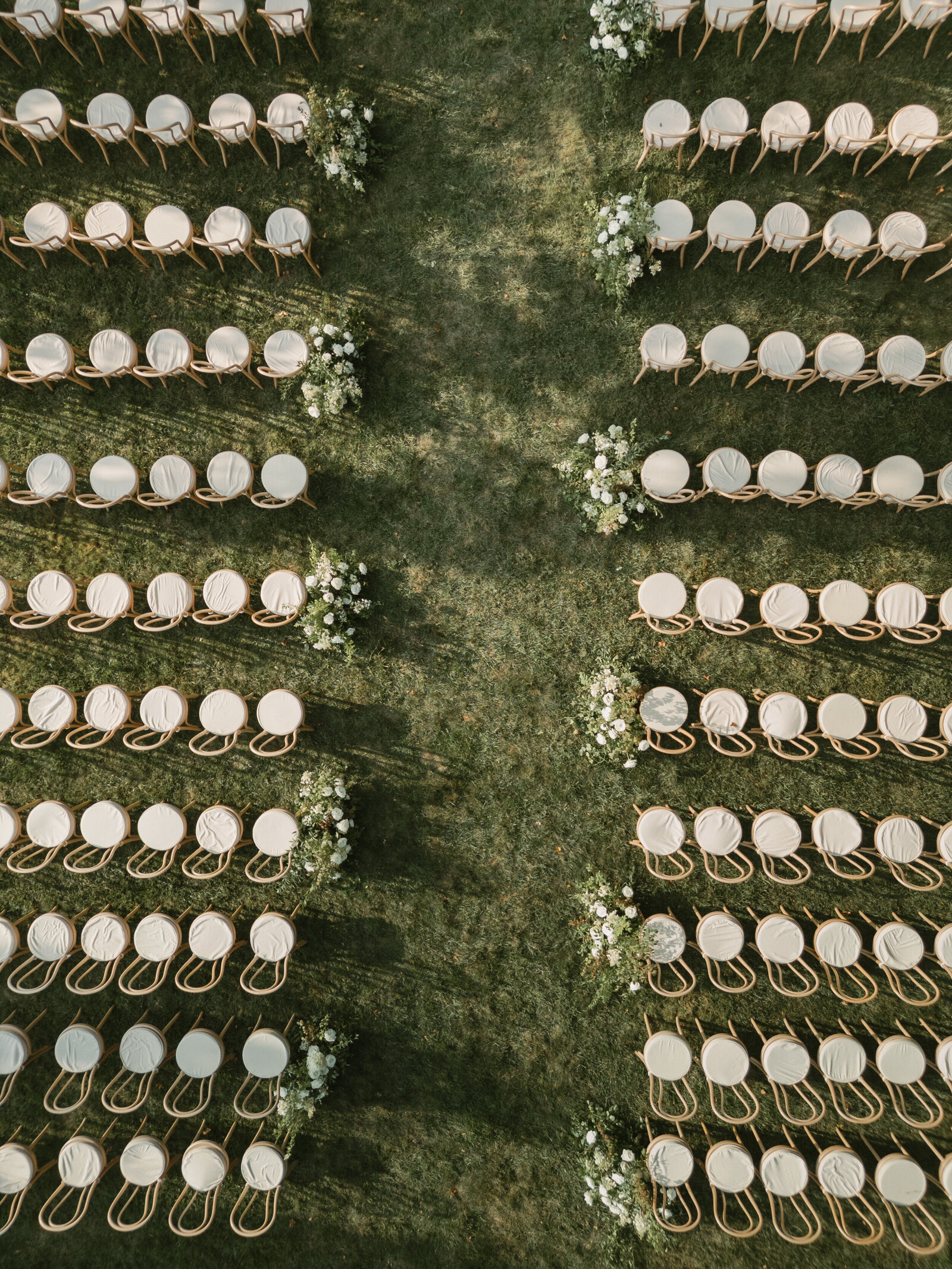 Overhead drone shot of white ceremony chairs with wooden backs lined up in rows and ceremony aisle running down the center lined in white floral arrangements 
