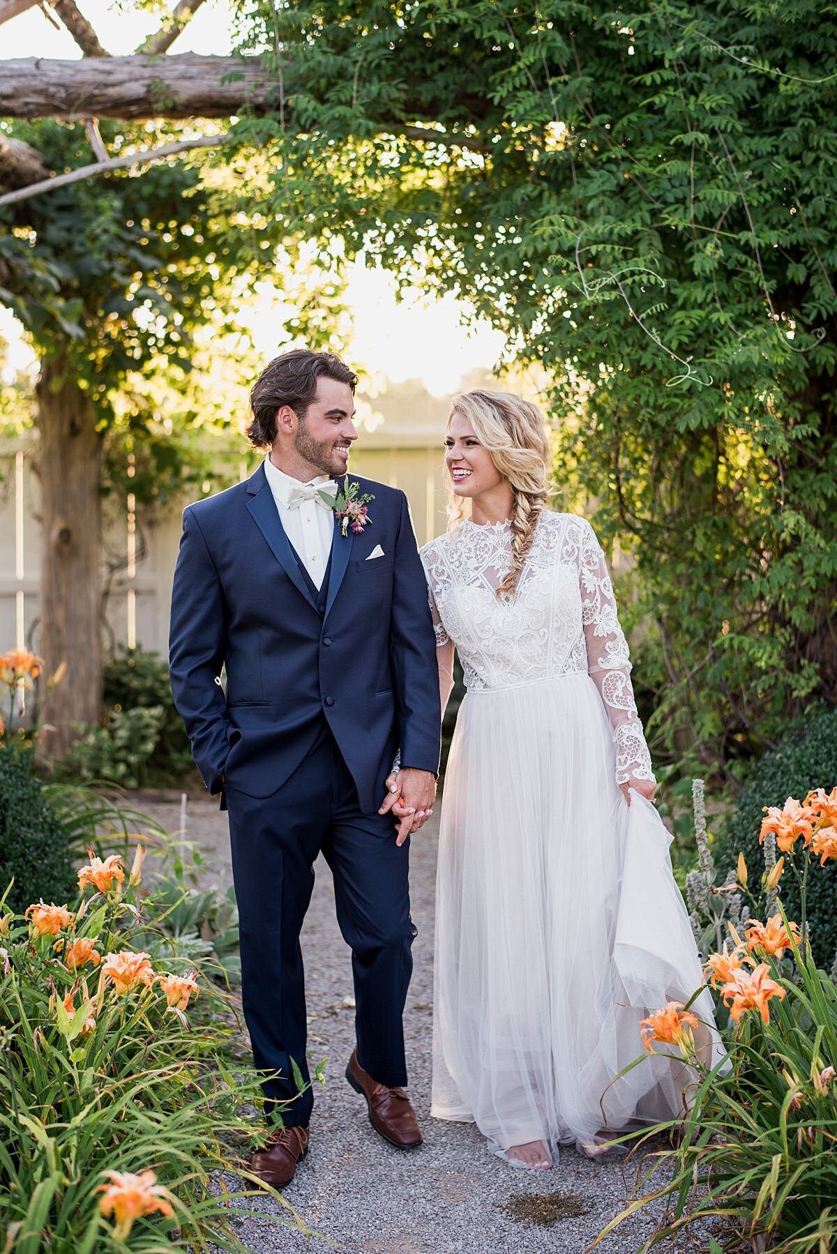 The groom, wearing a navy suit and mauve boutonniere leads the bride, wearing a boho lace gown through the garden filled with orange lilies