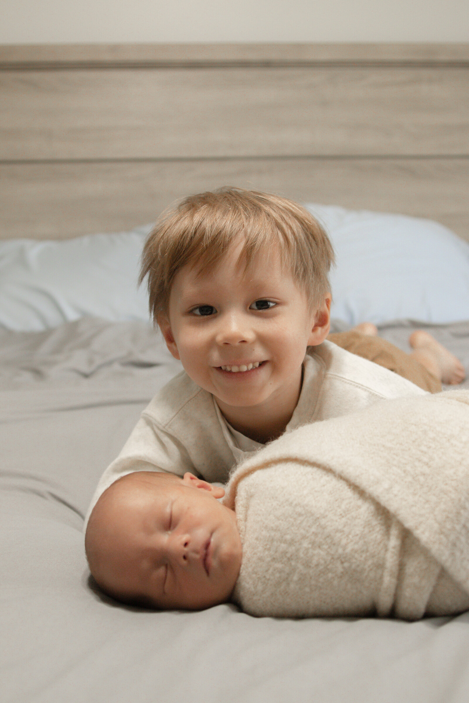 big brother smiles while laying on bed with little brother swaddled in front of him