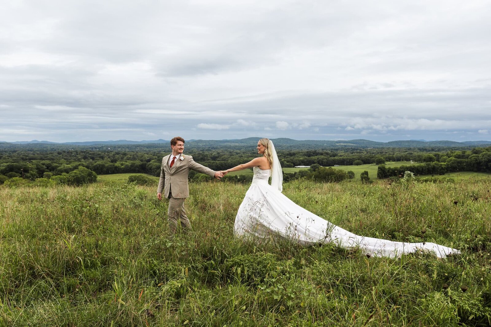 A bride and groom hold hands and walk through a wide-open grassy field, surrounded by rolling hills under a moody, overcast sky. The bride’s long train trails softly behind her as she looks back at her partner with a serene smile. The groom gently leads the way, dressed in a light brown suit and rust-toned tie. The moment feels grounded and intimate, like time has slowed just for them.