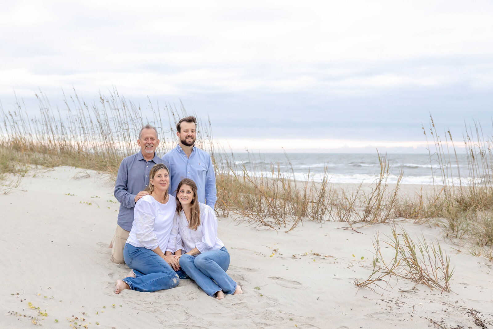  A family of four sits comfortably on the sandy dunes of Oak Island, dressed in coordinating blue and white attire. The overcast sky and gentle sea oats add a peaceful ambiance. This image highlights the best family photographer in North Carolina, offering artistic family photography in Wilmington, NC.