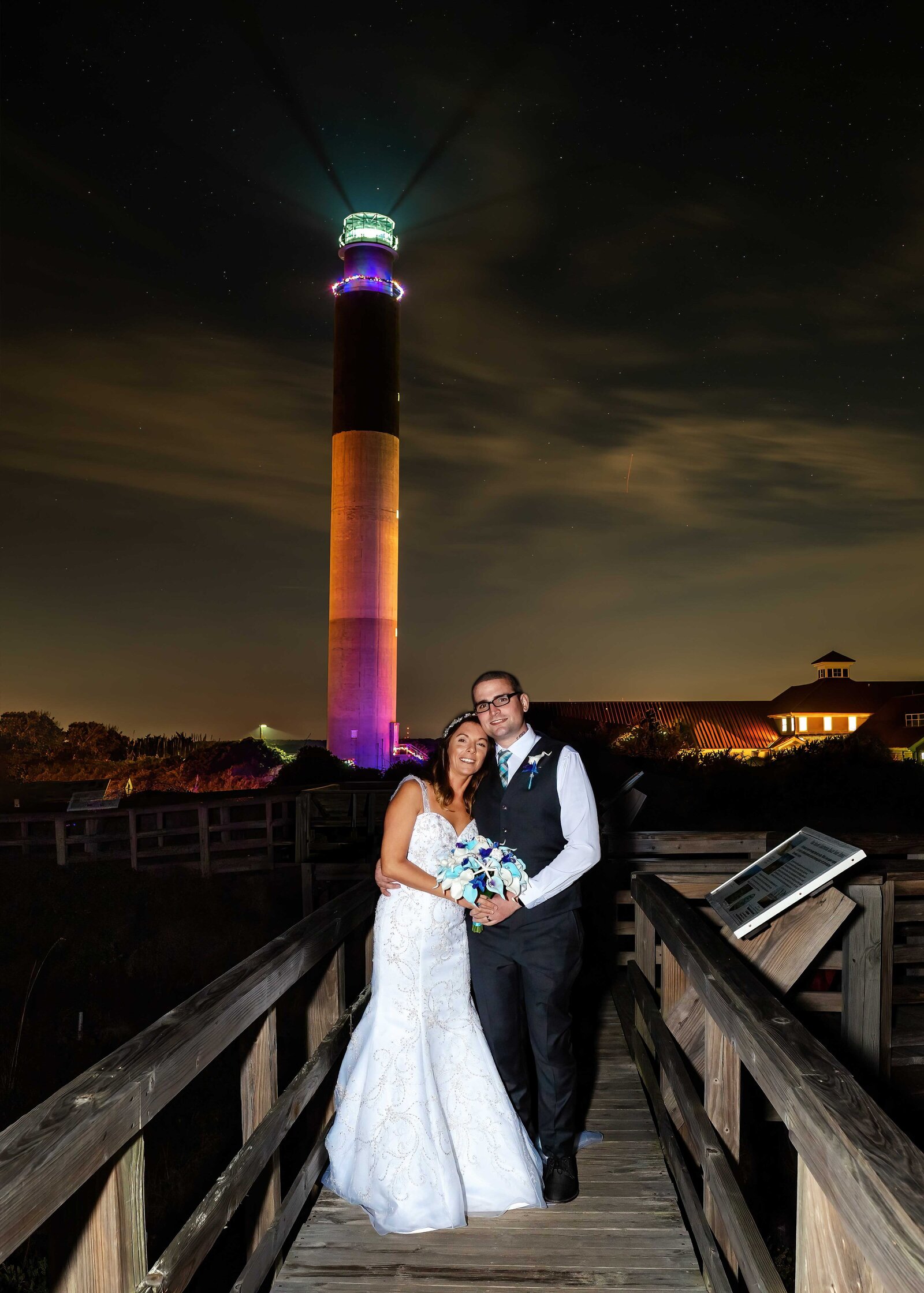 The bride and groom pose under a glowing Oak Island Lighthouse at night, captured on a wooden boardwalk during a Bald Head Island, NC wedding, blending coastal charm with romantic elegance.
