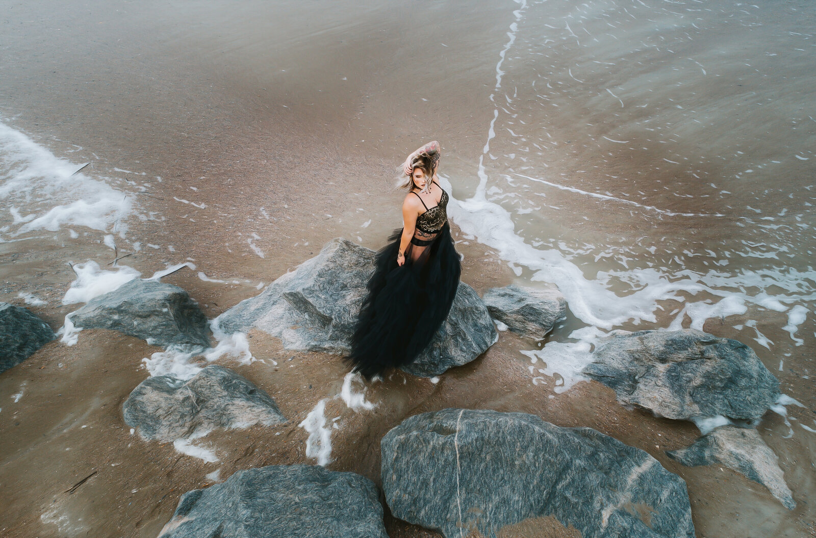 Aerial shot of woman on beach with black gown  standing on rocks