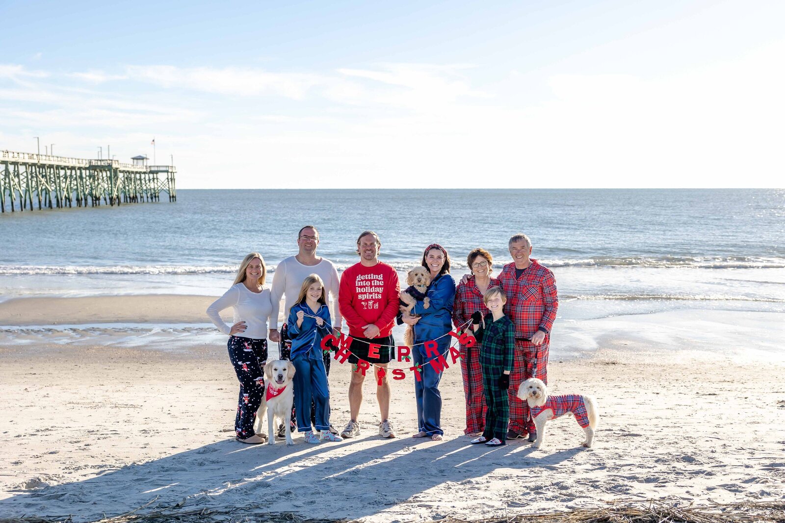 A family poses with their dogs on the beach, dressed in festive holiday pajamas. The ocean and pier in the background add to the cheerful scene, ideal for destination family photographer NC, capturing holiday joy by the sea.
