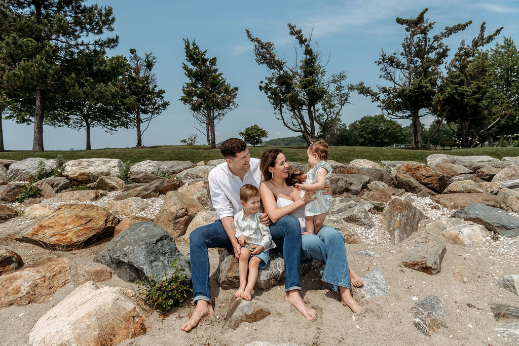 Connecticut Family Photographer captures an intimate moment of parents sitting on coastal rocks with their two young children, surrounded by natural stone formations and green trees