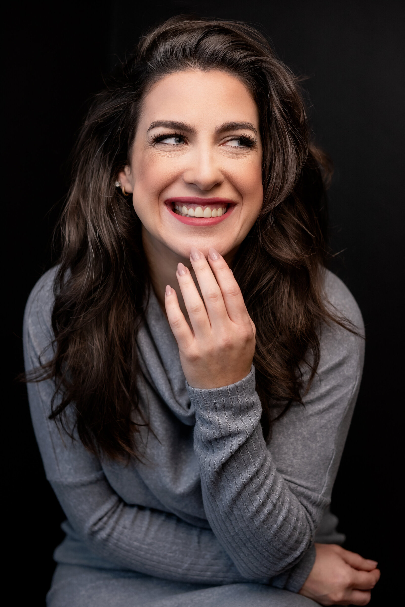 Headshot of a woman with long brown hair wearing a grey sweater dress smiling as she looks off-camera