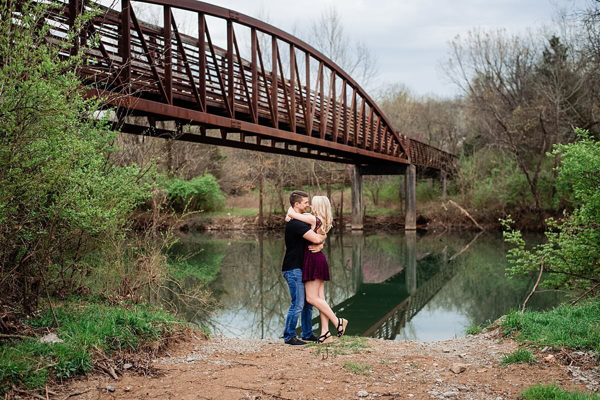 Engagement photo of couple hugging  next to a river with a bridge overhead on Murfreesboro greenway
